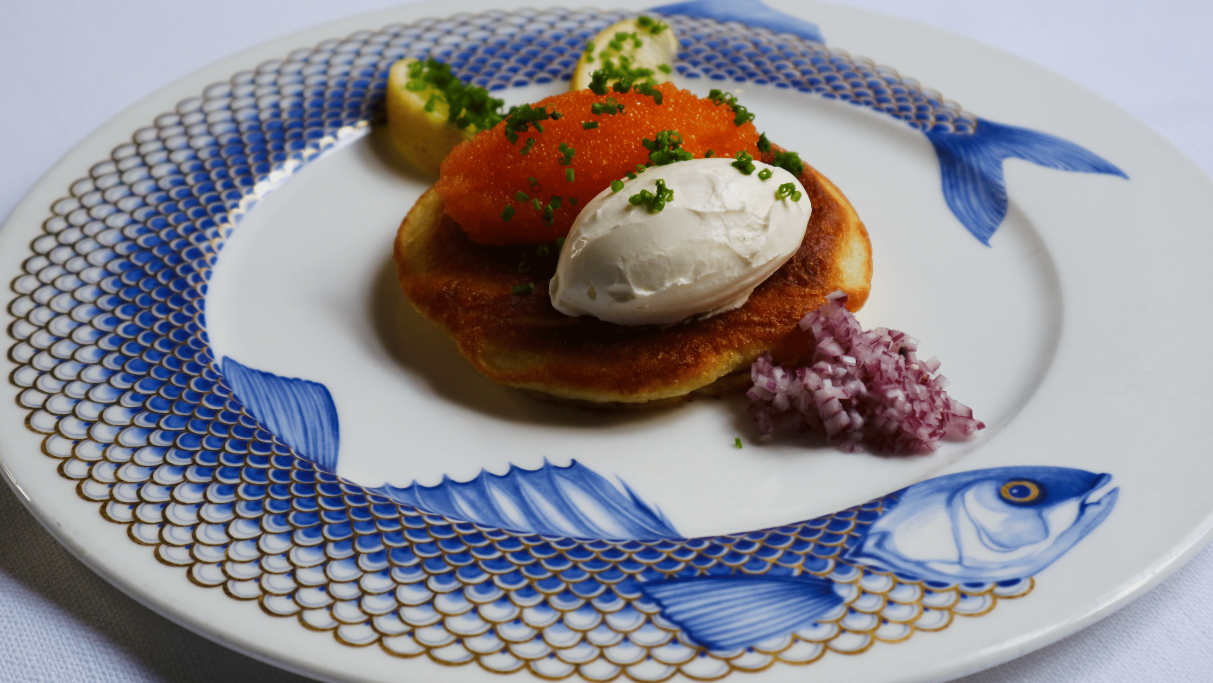 Fried fish fillet topped with sour cream, garnished with chopped chives, served with lemon wedges, red caviar, and diced red onion on a decorative fish-patterned plate.