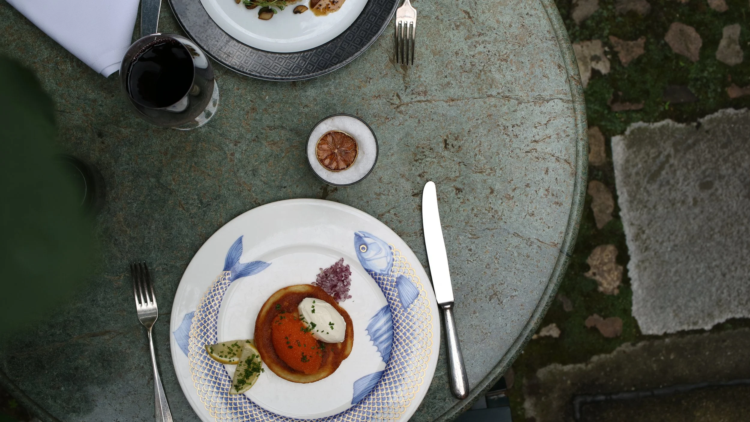 A table set outdoors with a plate of food, a glass of red wine, and a lemon slice. The plate includes a serving of food with garnishes. The table has a rustic, weathered surface.