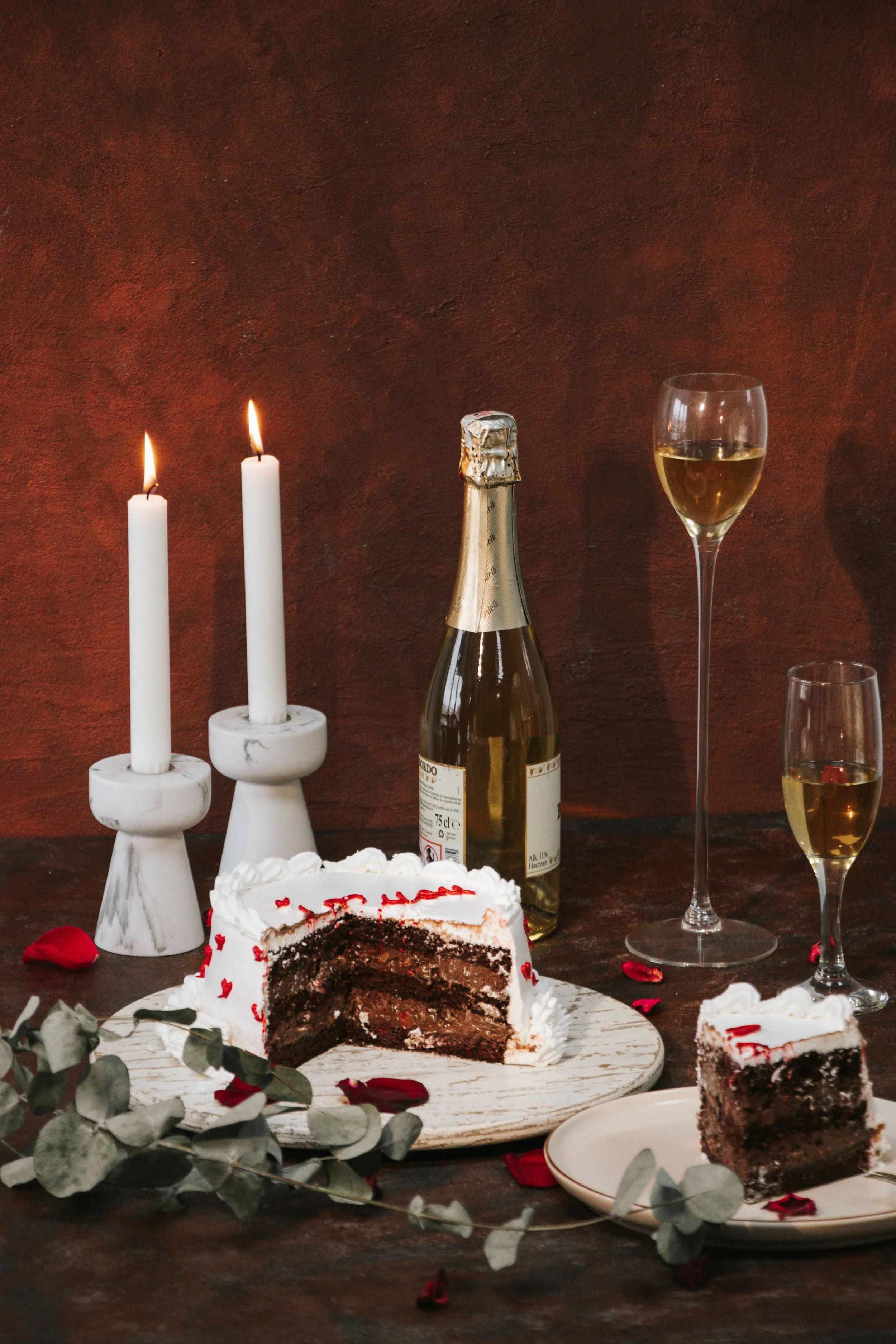A partially sliced chocolate cake with white frosting and red heart-shaped decorations, on a white platter, surrounded by rose petals and eucalyptus leaves, with a bottle of champagne, two glasses of white wine, white candles, and a cake with red writing in the background.