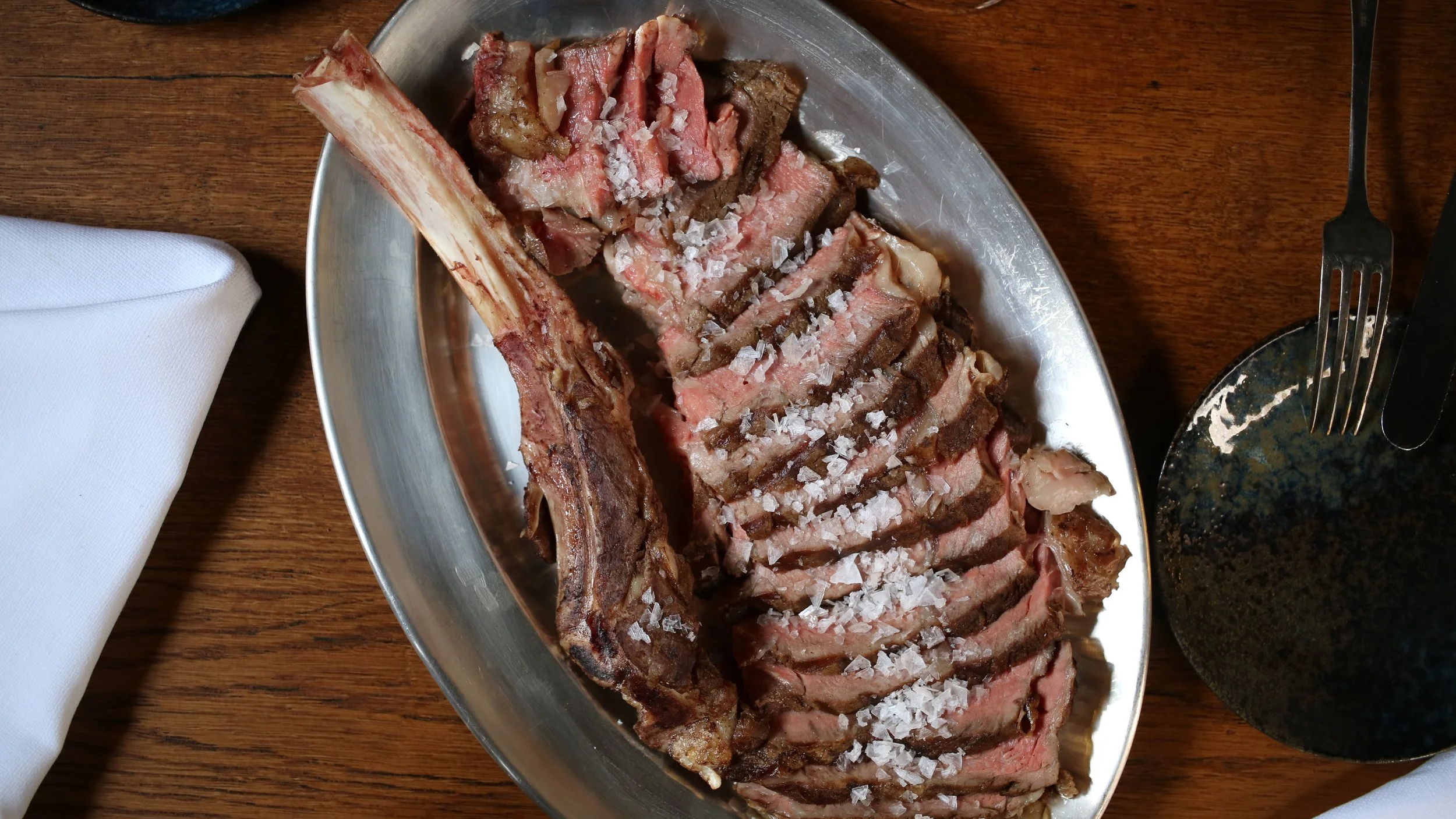Sliced cooked steak with sea salt on a metal serving platter on a wooden table.