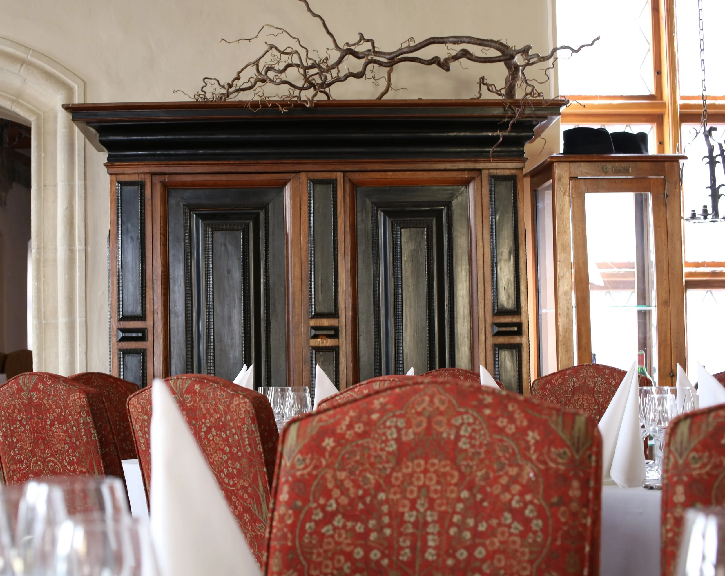 Elegant dining room with red patterned chairs, white tablecloths, and glassware, with a large wooden cabinet and decorative branch on the wall.