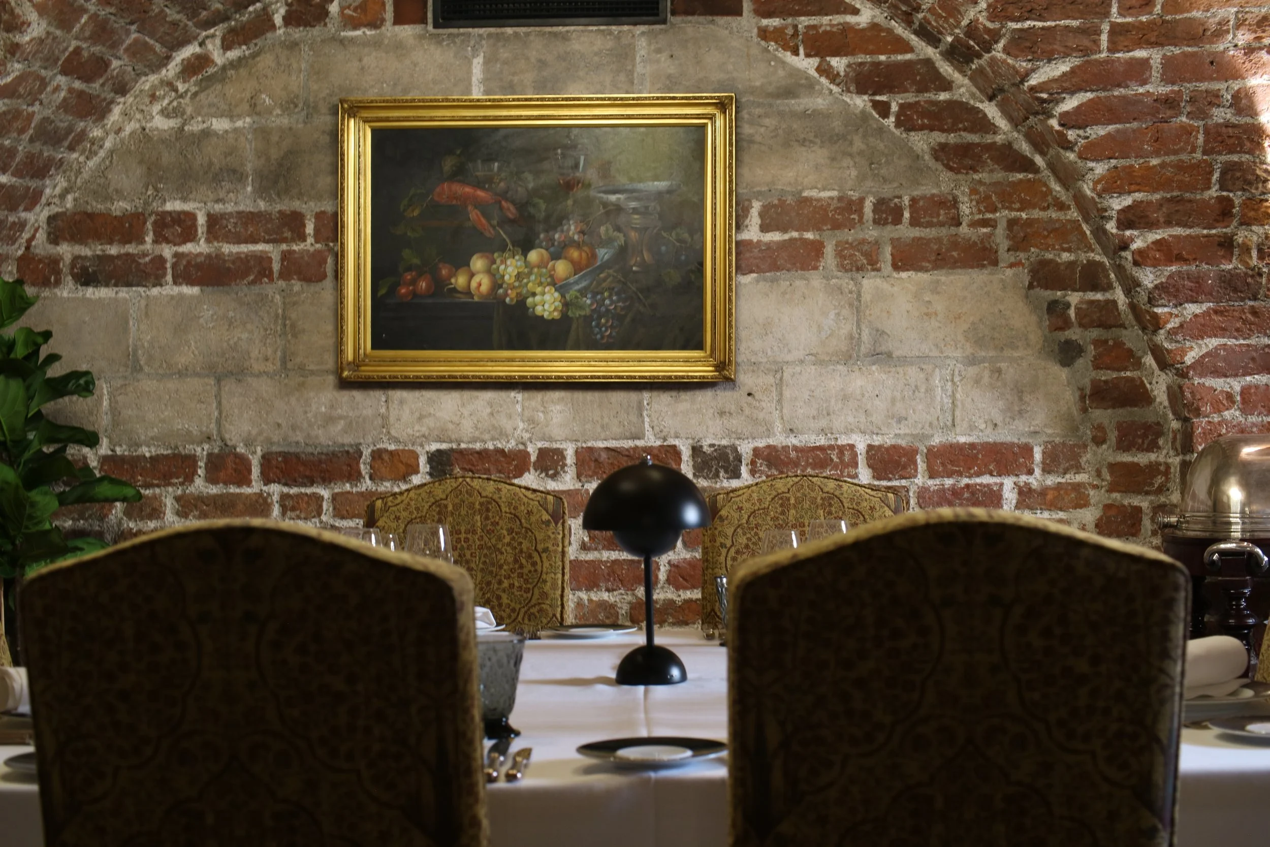 A dining room with a brick and concrete wall, a framed still life painting of fruit, a table set for dining, and upholstered chairs.