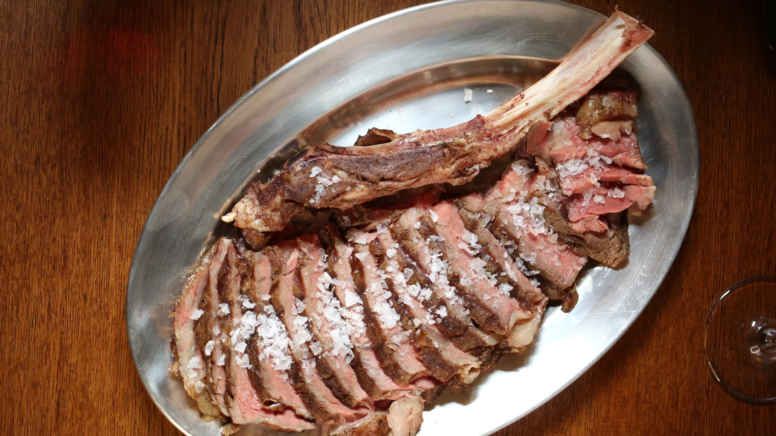 Sliced cooked T-bone steak seasoned with salt on a metal platter on a wooden table.