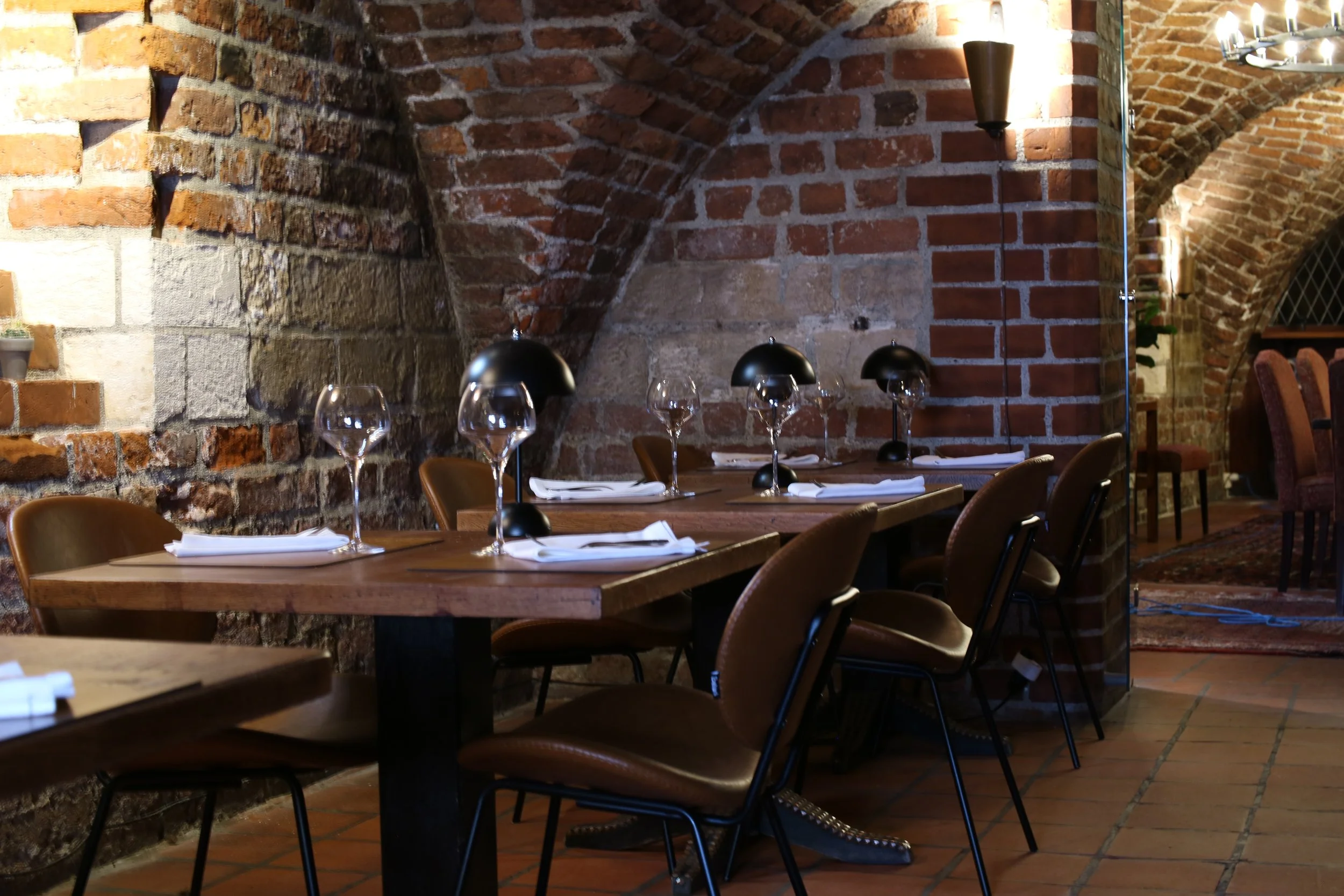 Dining area with wooden tables set with white napkins, wine glasses, and black lamps, against exposed brick walls with arched ceilings and lighting fixtures.