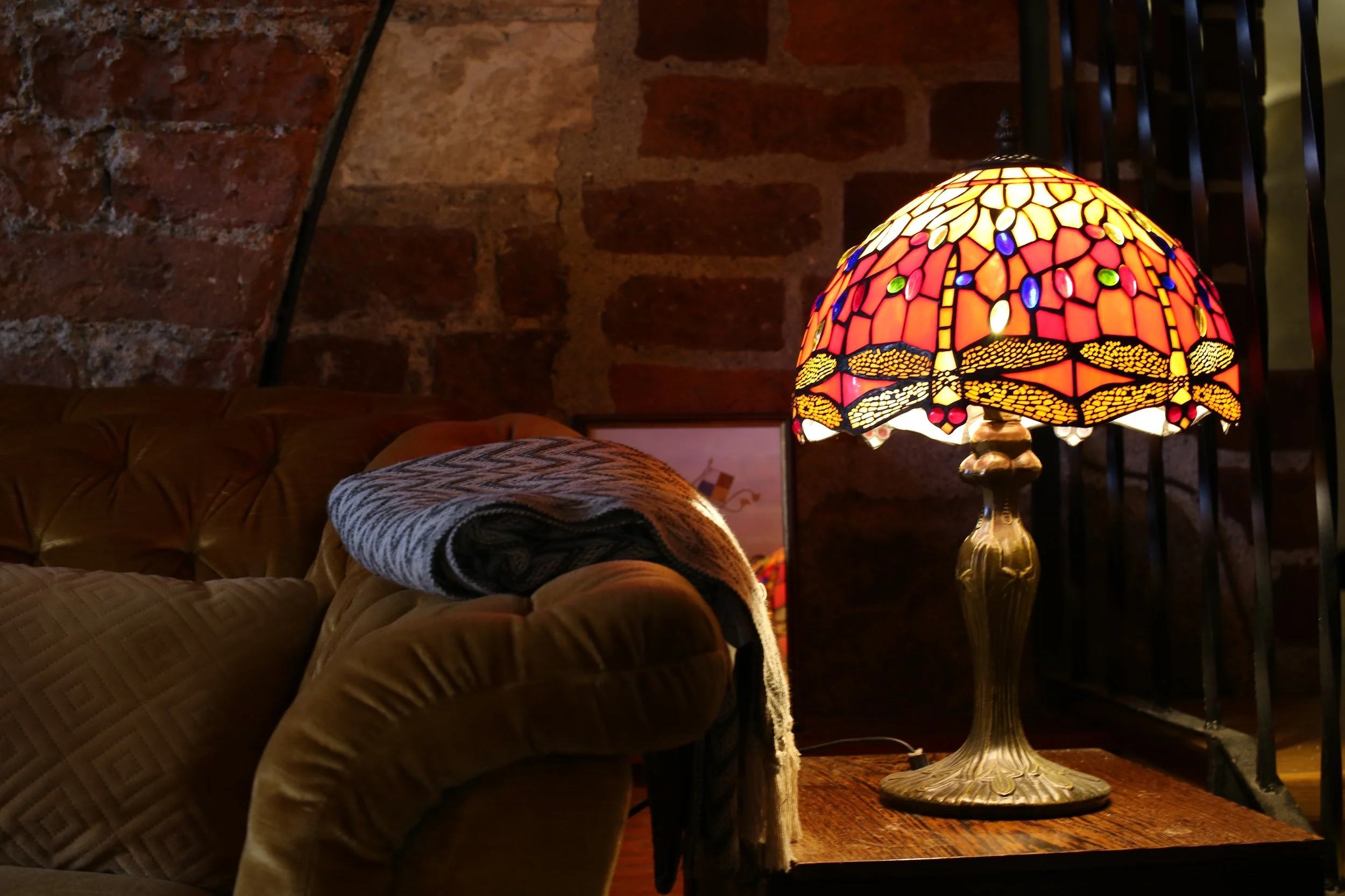 A Tiffany-style stained glass table lamp with a dragonfly design on a wooden side table, in a dimly lit room with a brick wall in the background, and a plaid blanket on a sofa.