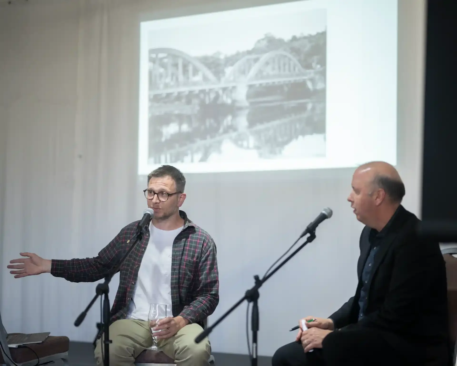 Two men sitting at a panel discussion or interview, with microphones in front of them. The man on the left is gesturing with his hand, holding a wine glass, and wearing glasses and a plaid shirt. The man on the right is looking at the speaker. A projector screen behind them displays an image of a bridge over a body of water.