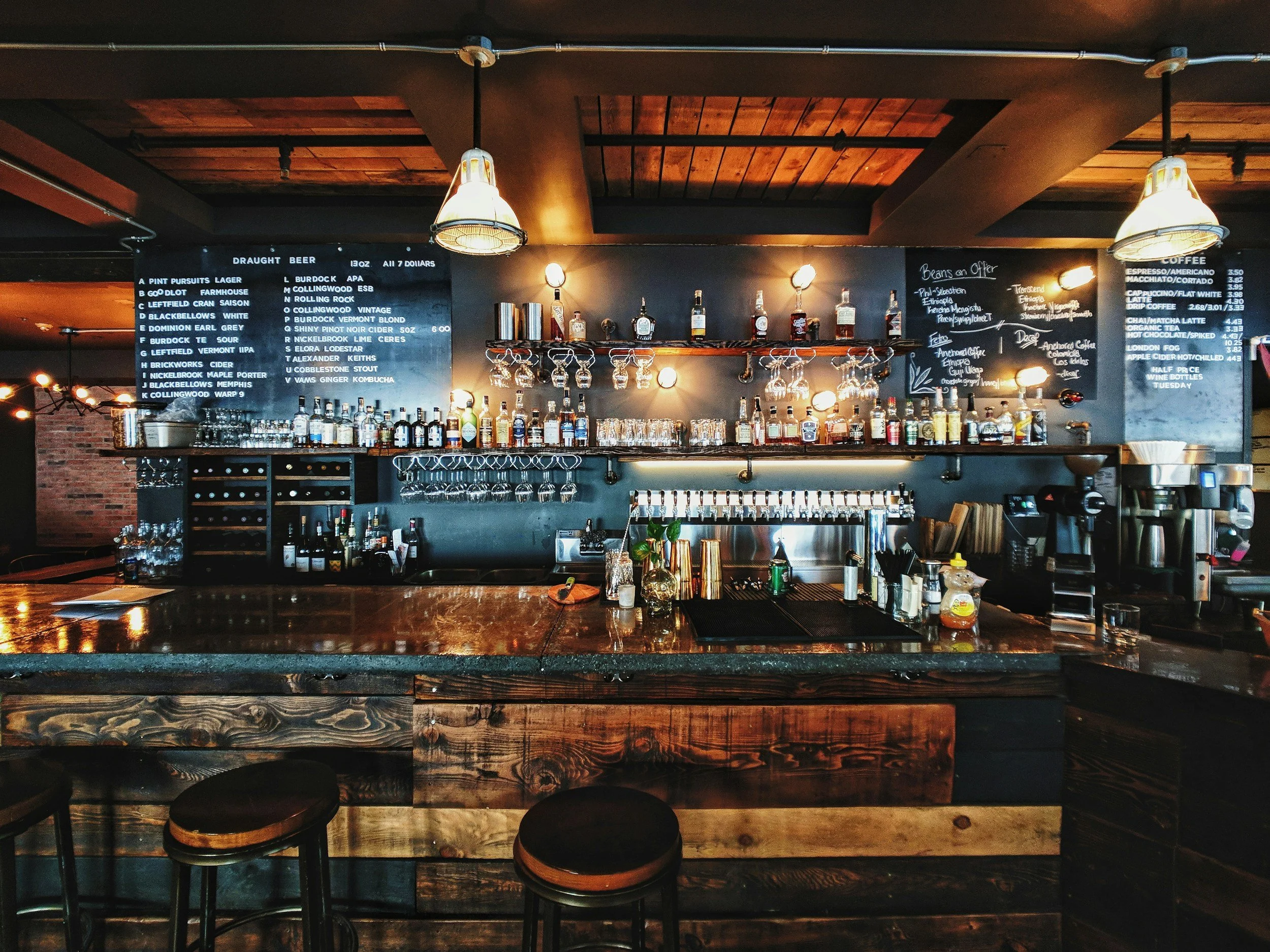 A bar interior with liquor bottles, hanging wine glasses, beer on tap, and menu boards, featuring warm wooden accents and black walls.