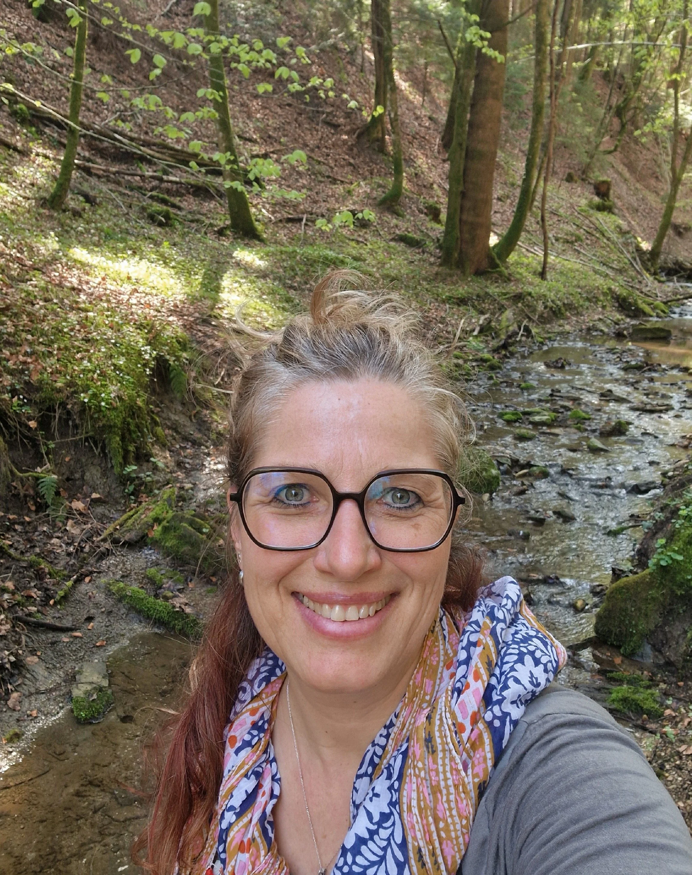 Eine lachende Frau - Monika Schimanski - mit Brille, lange Haare, in einem Wald an einem Bach, im Hintergrund Bäume und Moos.