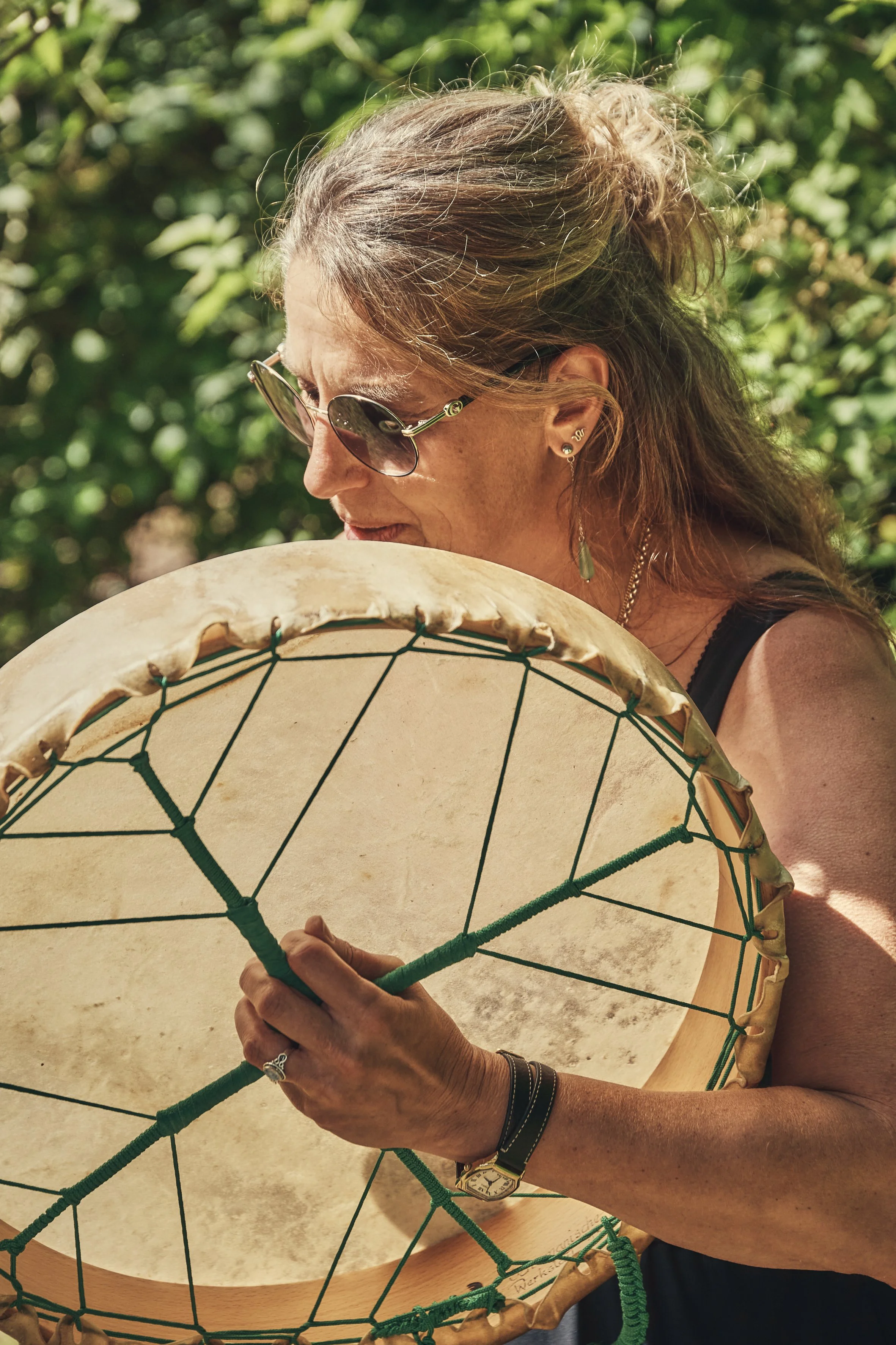 Eine Frau - Monika Schimanski - mit brünettem Haar trägt eine Sonnenbrille und Ohrringe und trommelt innig bei einem Ritual mit einer Schamanischen Trommel.