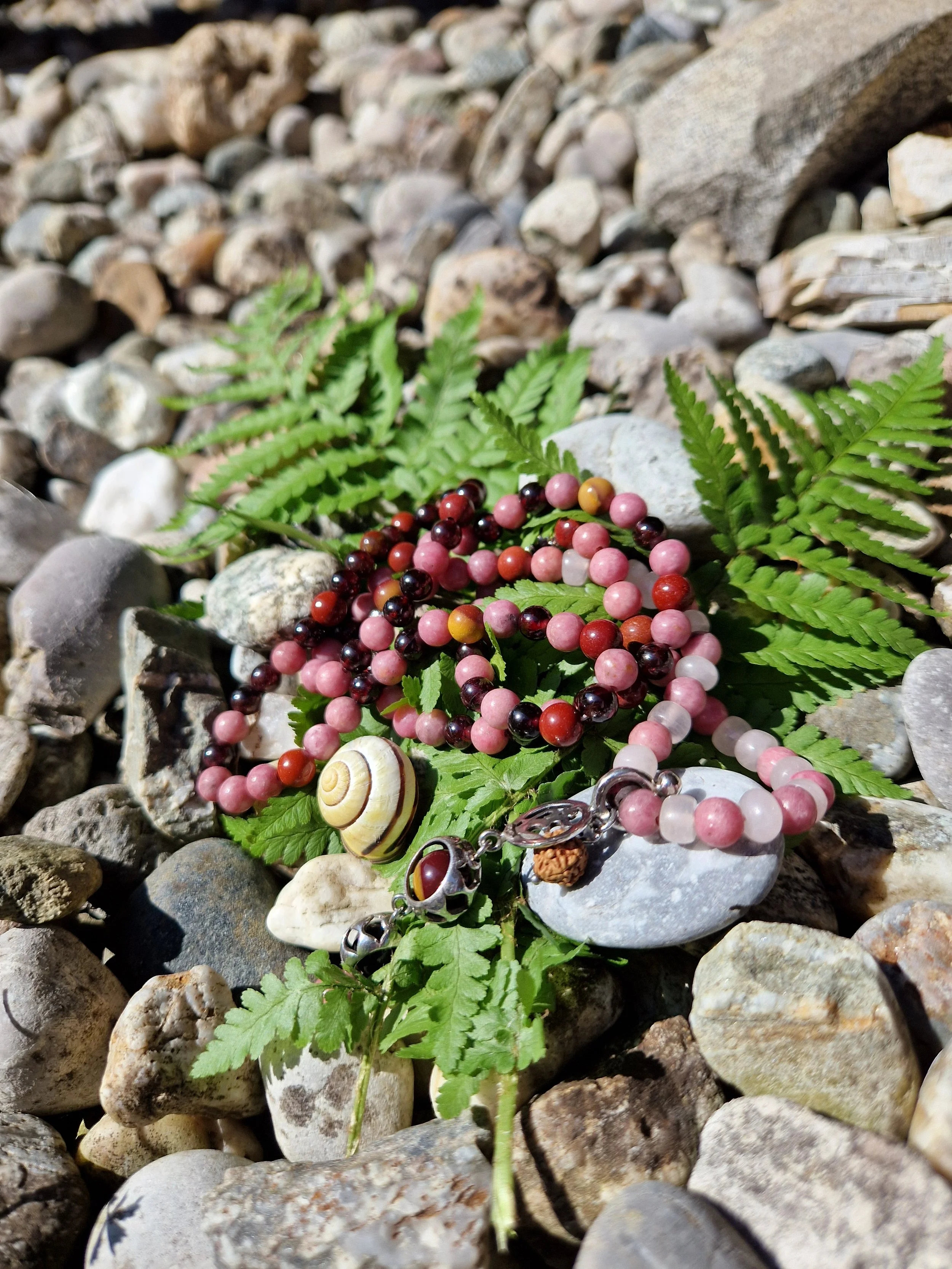 Eine Medizin-Halskette mit bunten Heilsteinen auf grünen Farnblättern auf einem Kieselsteinboden im Sonnenlicht.