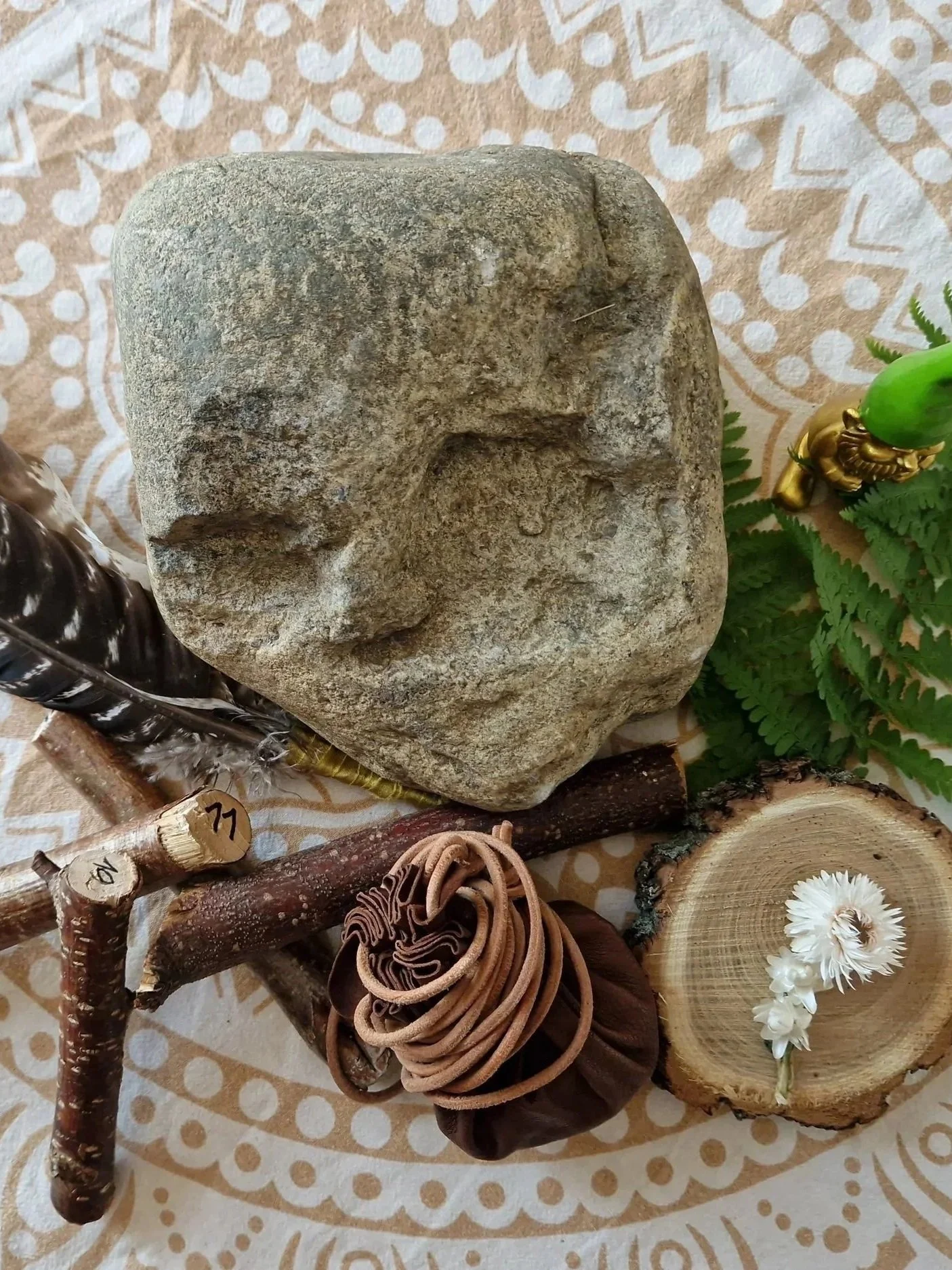 Stein mit mystischem Gesichter, umgeben von einem Schamanischen Medizinbeutel, Holzstücken, Pflanzen und einer weißen Blume auf einer gemusterten Ritualdecke.