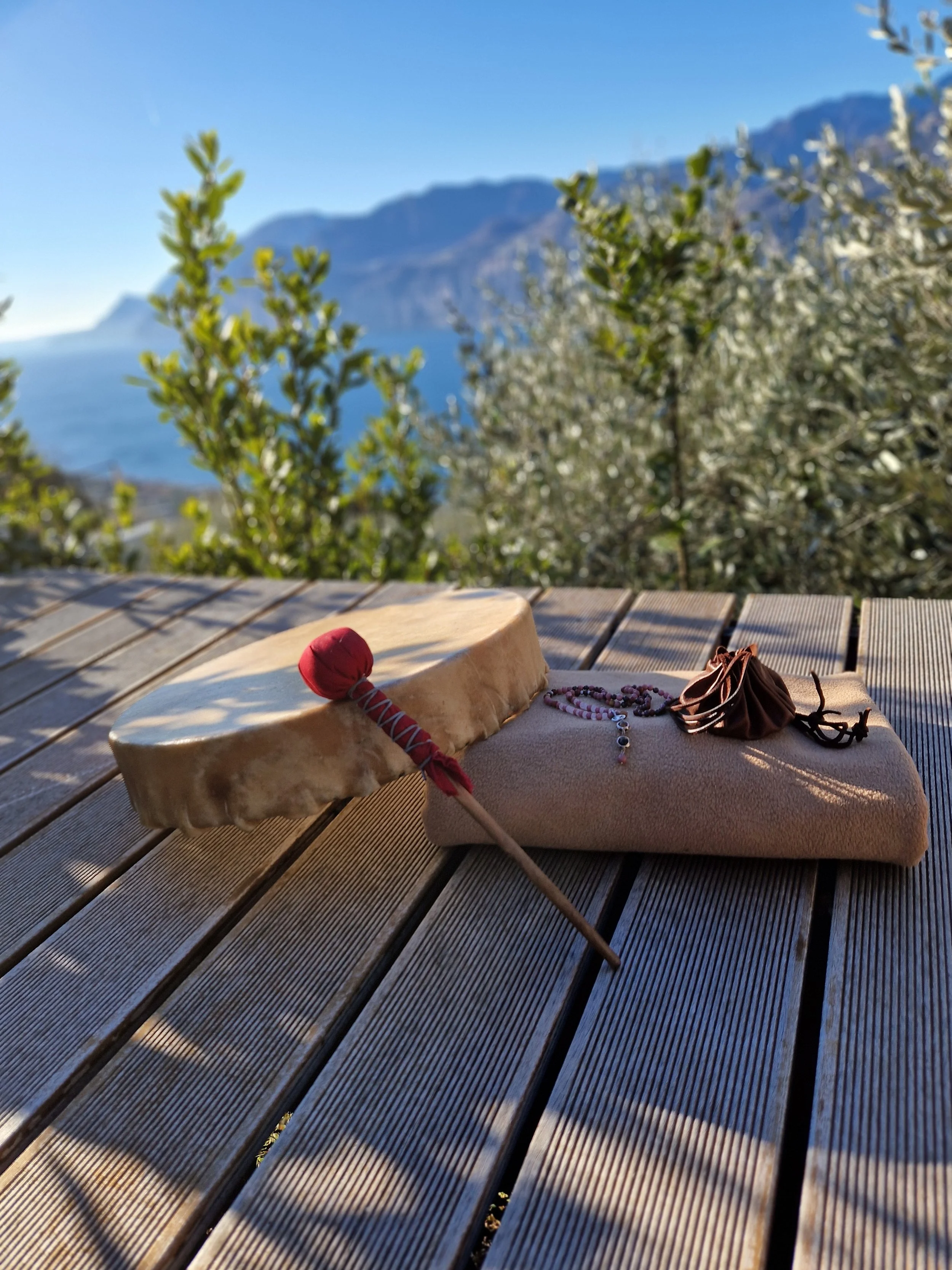 Auf einer Holzterrasse liegen eine Schamanische Trommel mit rotem Schlägel, eine braune Ritualdecke und ein Schamanischer Medizinbeutel. Im Hintergrund sind Berge, blauer Himmel und blühende Bäume sichtbar.