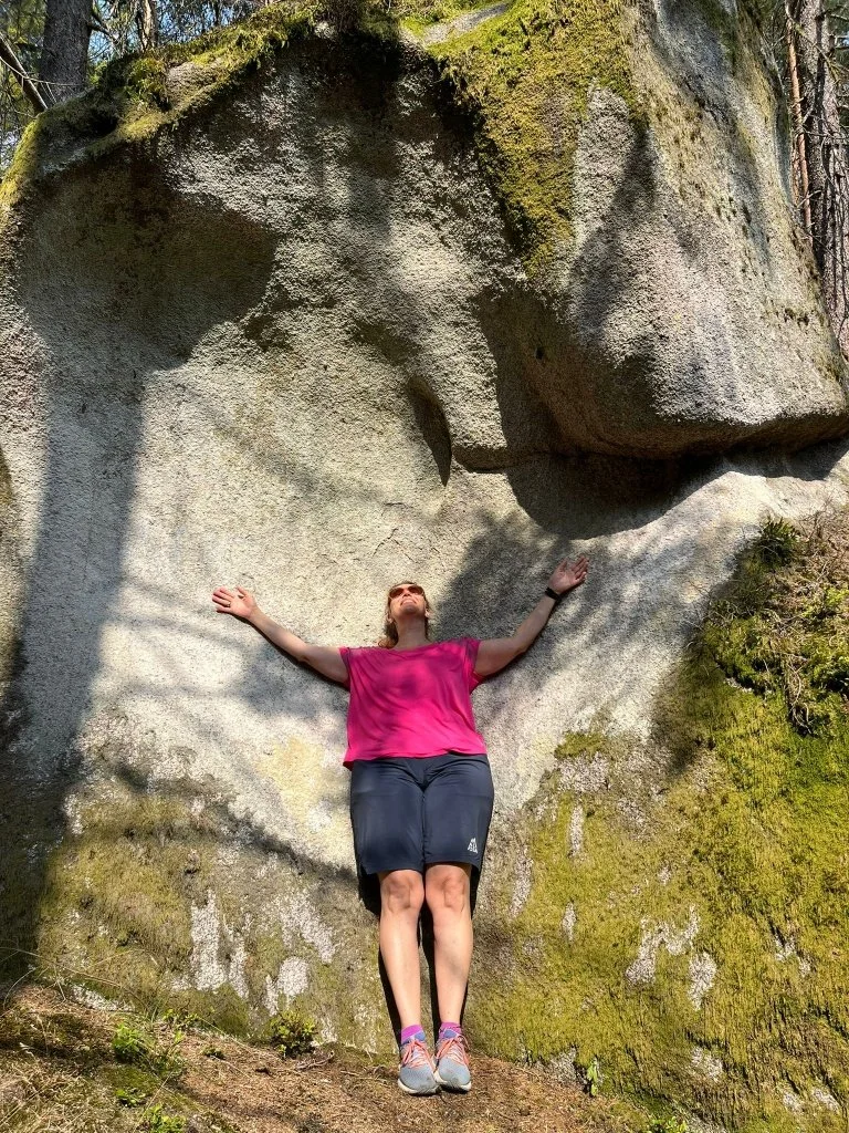 Eine Frau in rosa T-Shirt und schwarzen Shorts steht mit ausgestreckten Armen vor einem großen Felsen im Wald, der eine Gesichtsstruktur aufweist.
