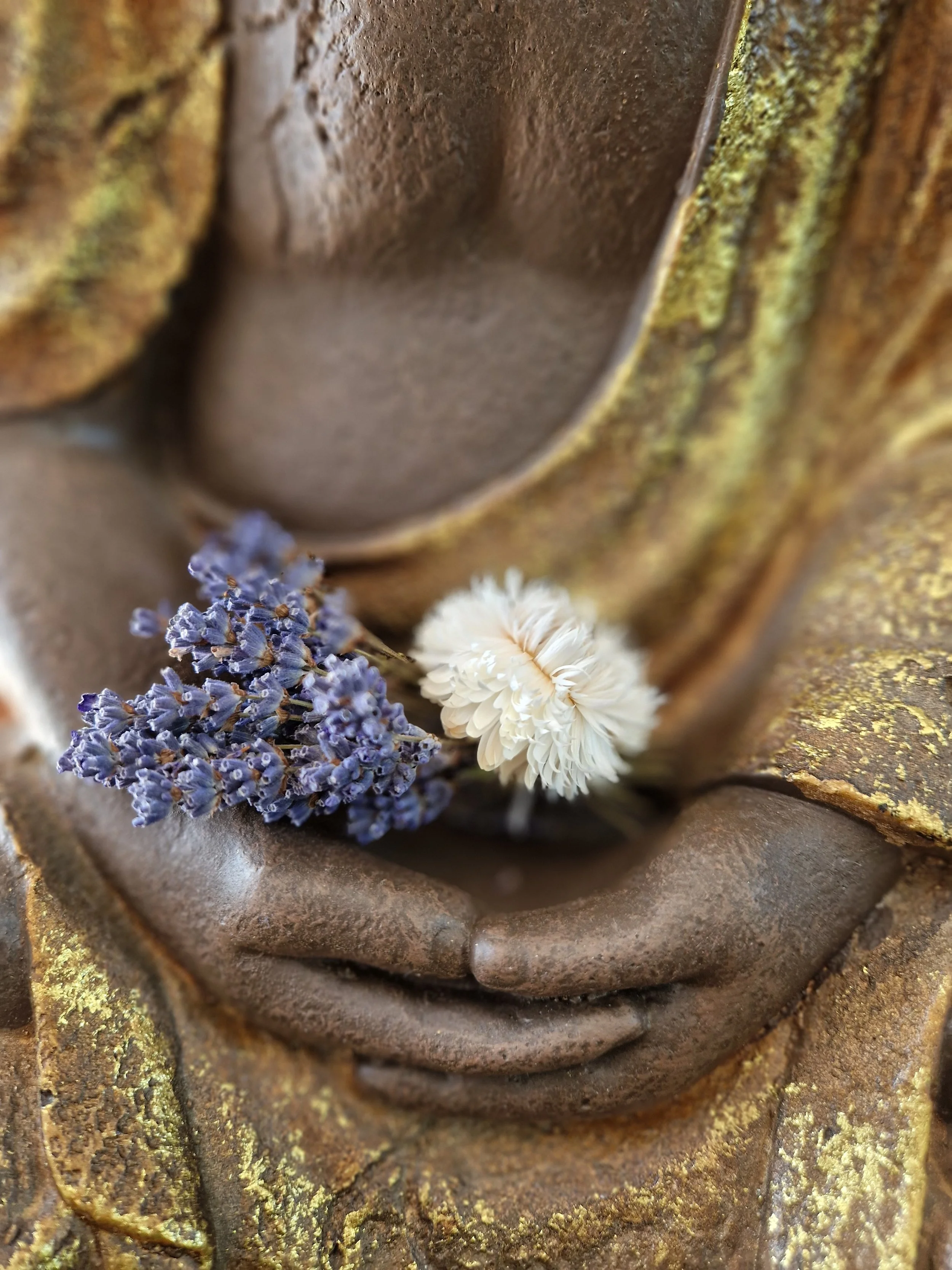 Nahaufnahme der Handhaltung einer goldenen Buddhafigur. Er hat ein getrocknetes Lavendelbündel und weiße Blumen im in der Hand.