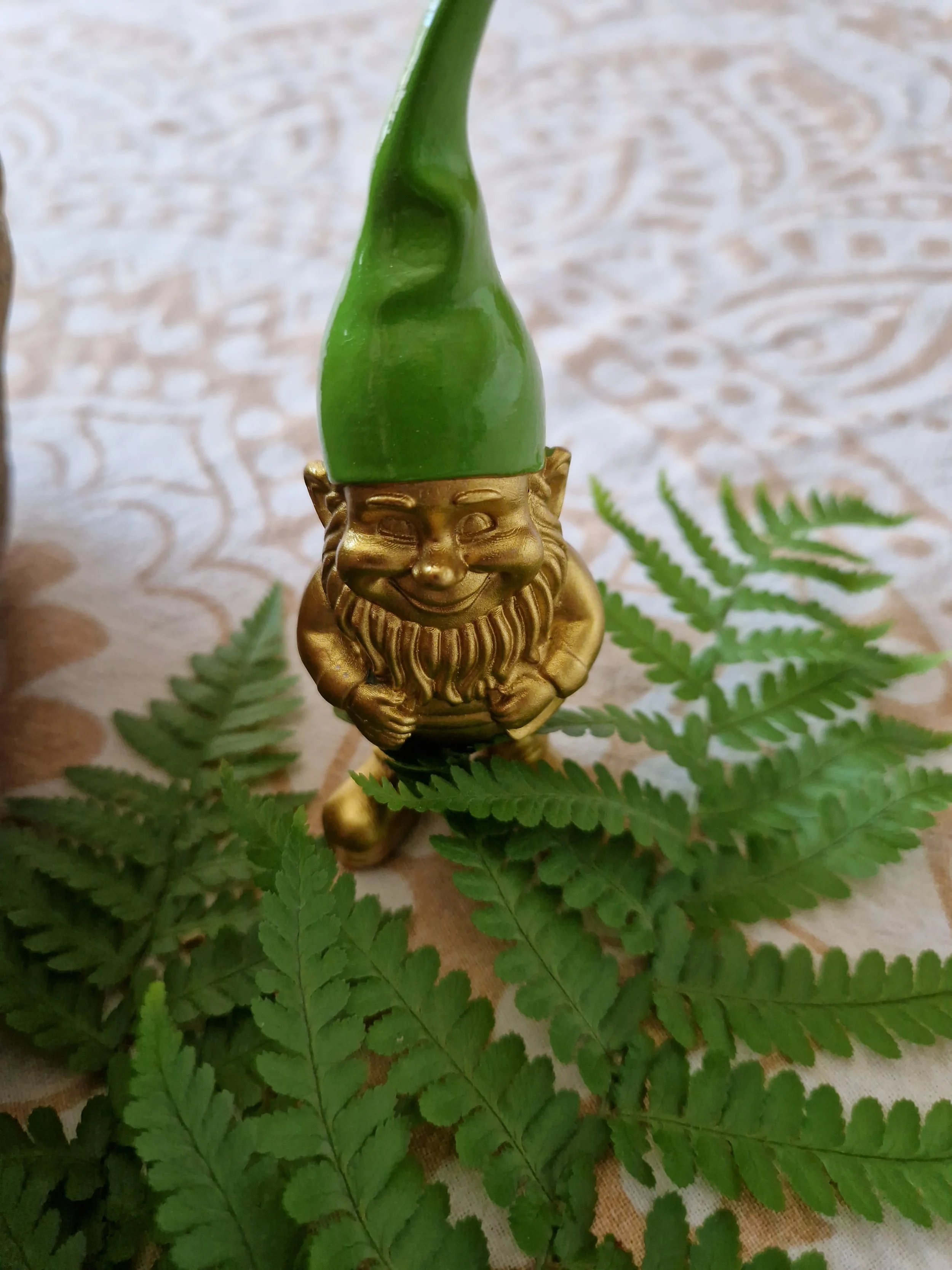 Goldener kleiner Deko-Zwerg mit einem grünen, spitzen Hut steht auf einer gemusterten Ritualdecke, umgeben von grünen Farnblättern.