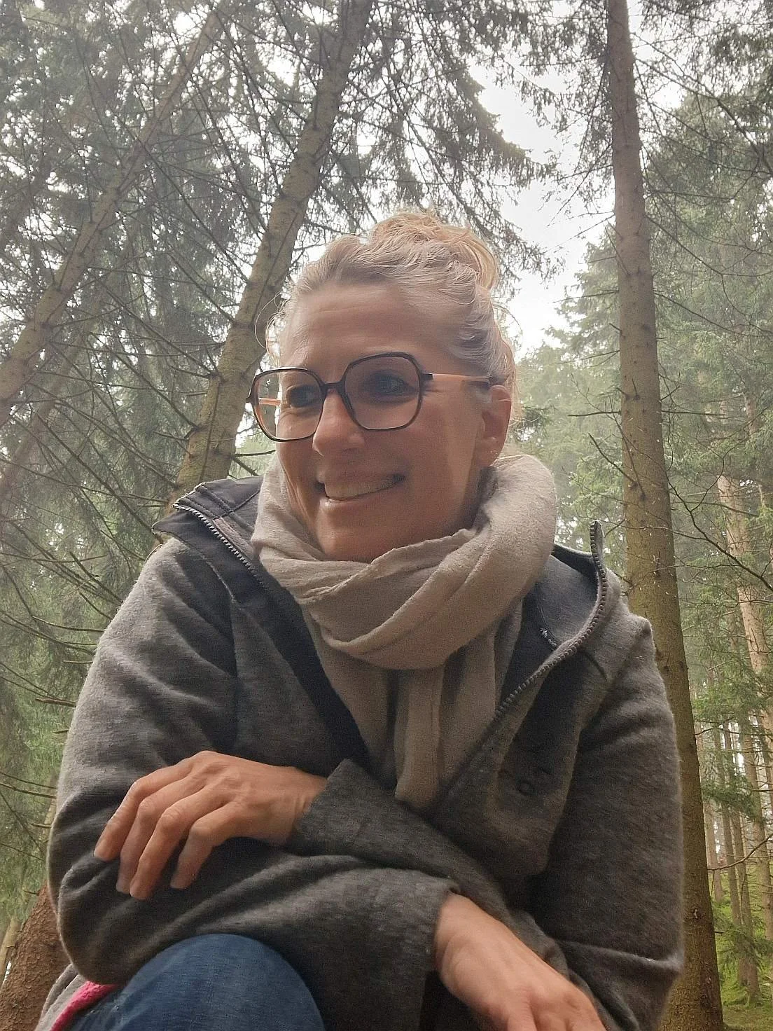 Eine lachende Frau - Monika Schimanski - mit Brille, in einem Wald, trägt eine graue Jacke und einen beige Schal.