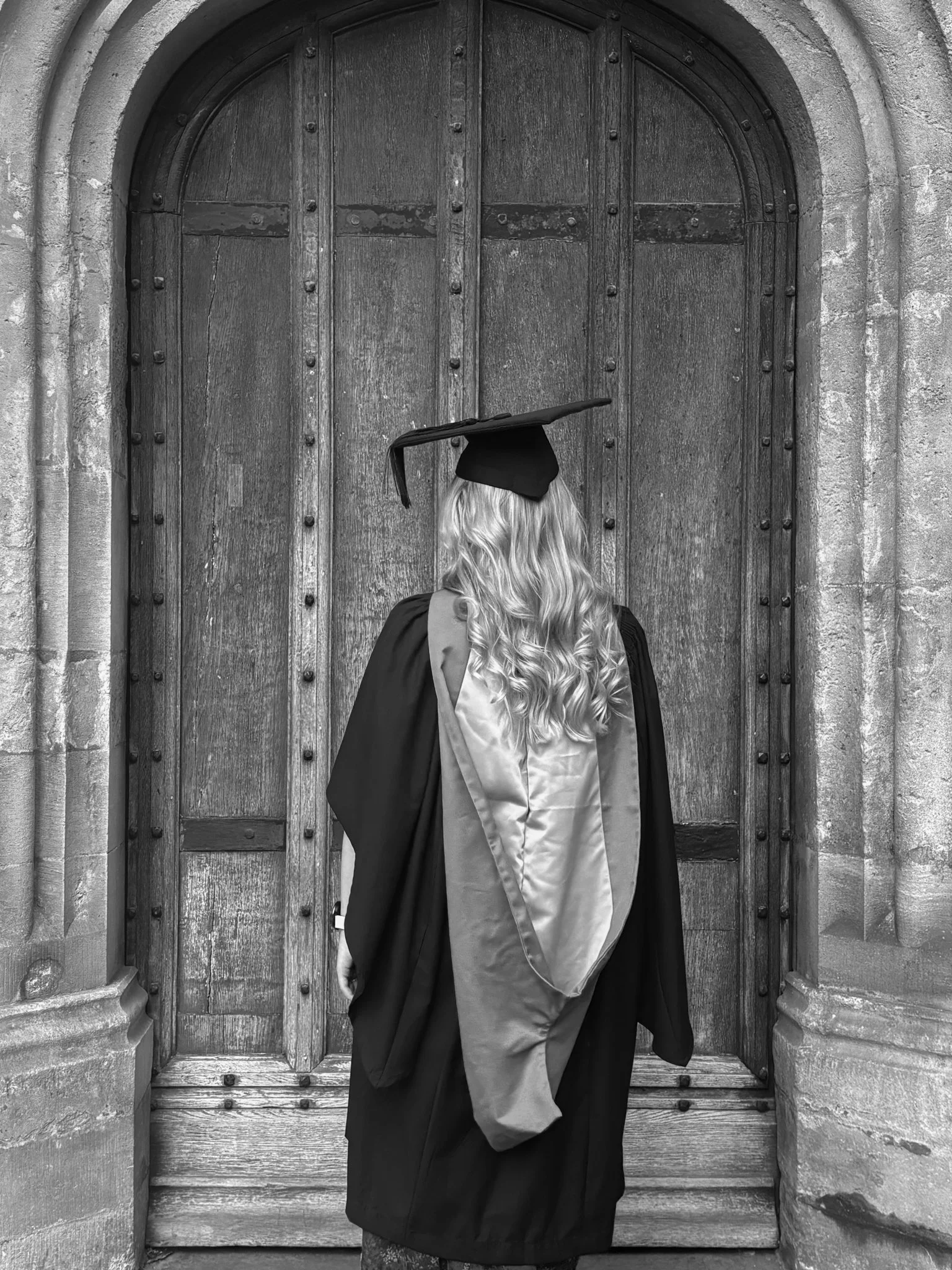 Person wearing graduation cap and gown standing in front of a large wooden arched door.