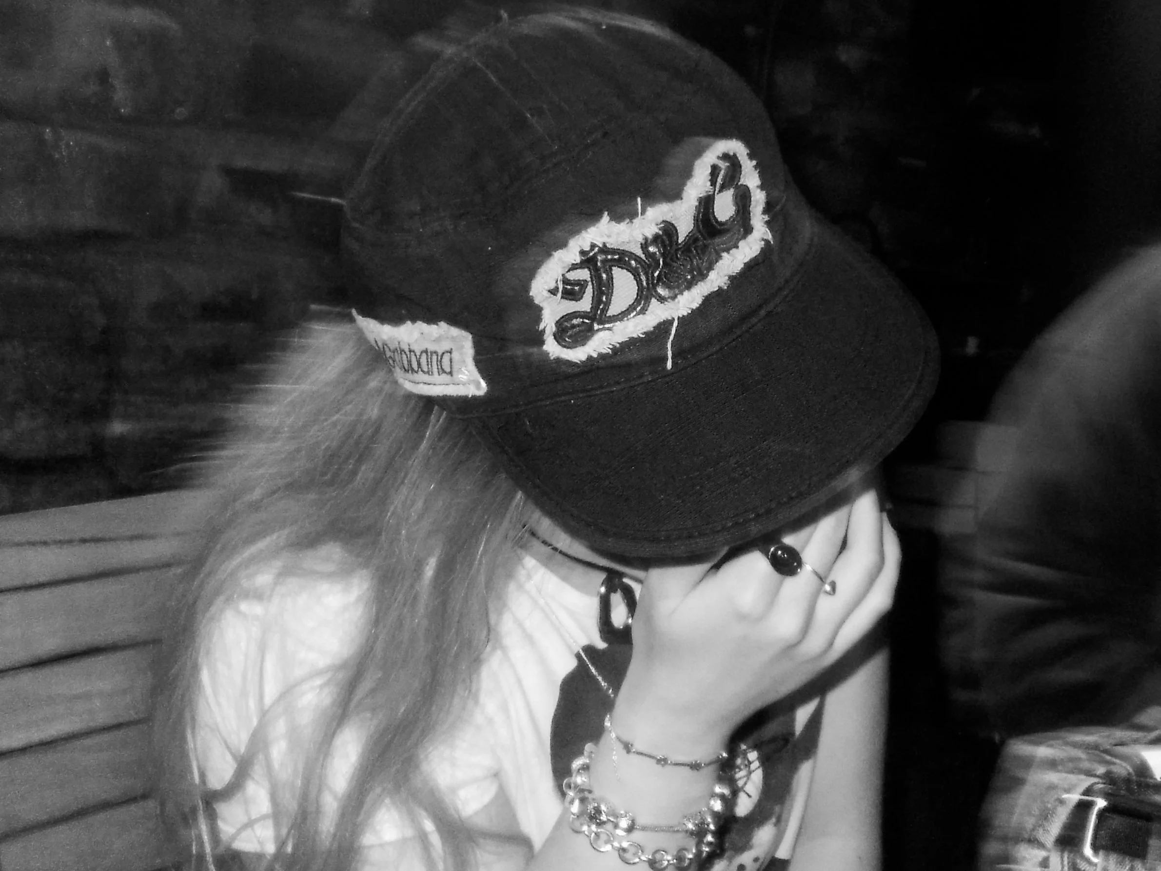 Black and white photo of a person wearing a baseball cap with the word 'Dior' embroidered on it, various jewelry, and a ring, with their hand resting on their face.