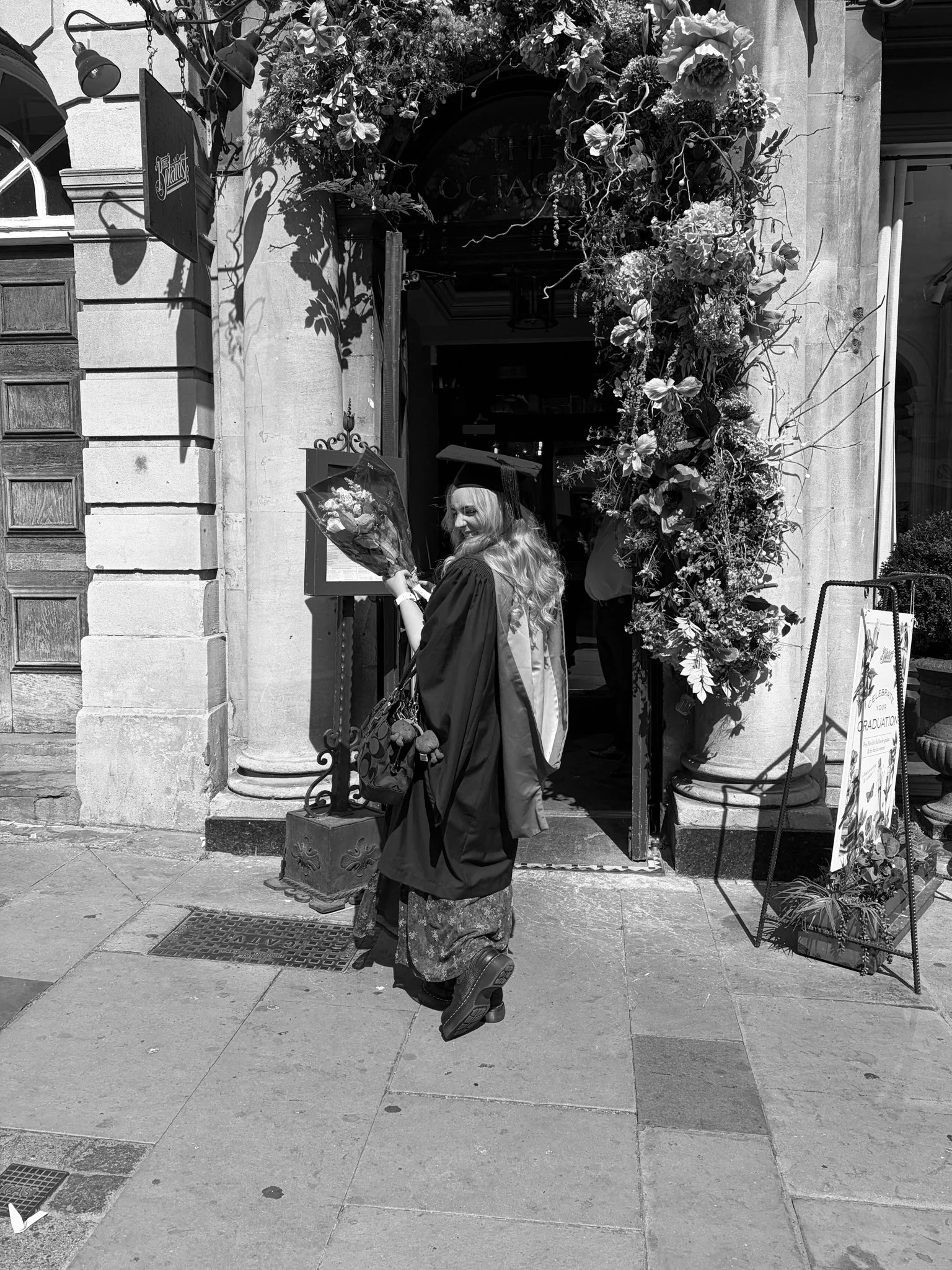 A woman in graduation cap and gown holding a bouquet of flowers outside a building decorated with flowers, with another person partially visible inside.