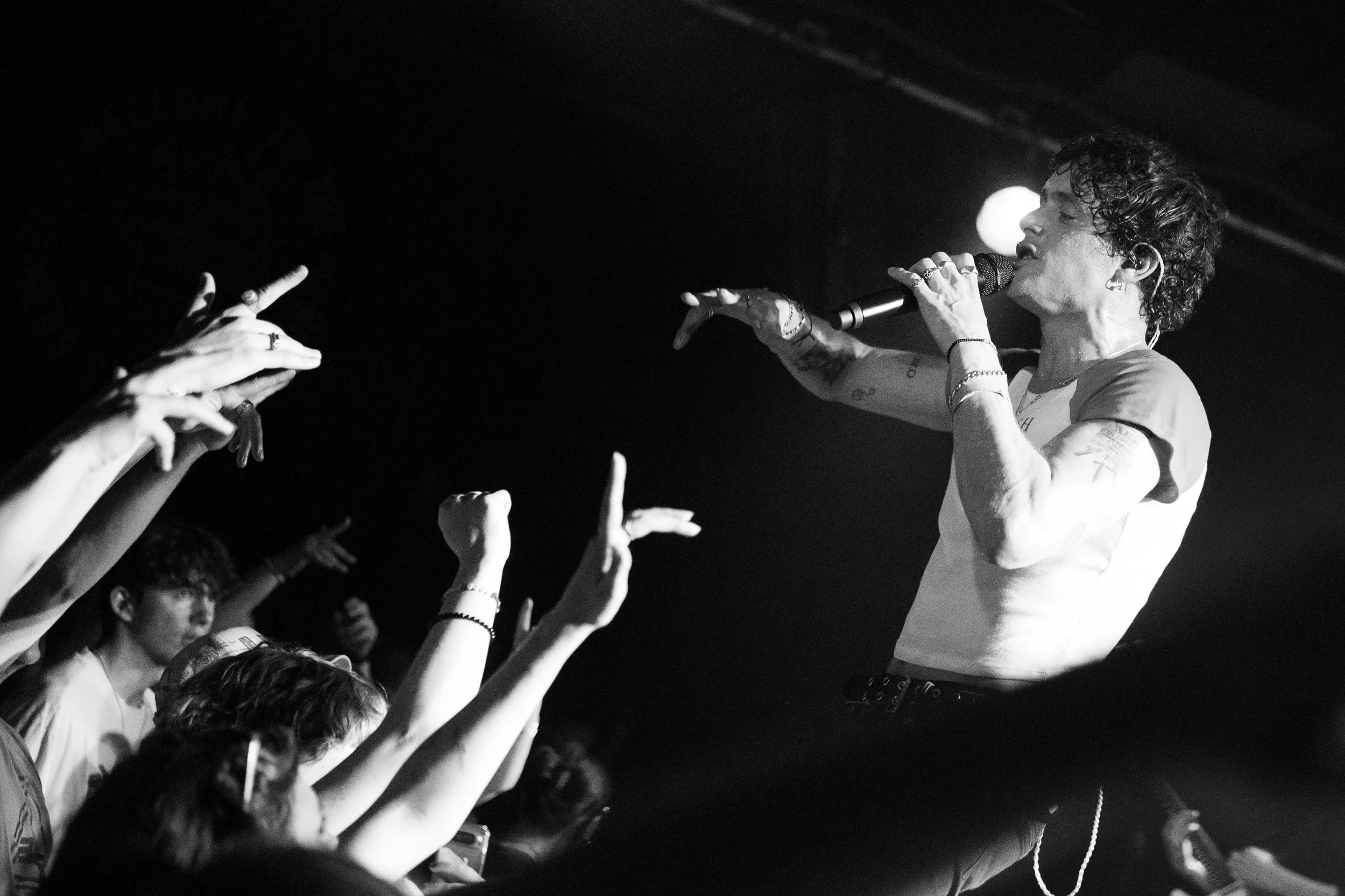 A singer with curly hair performs on stage, holding a microphone, while reaching out to fans with their hand in a crowded concert.