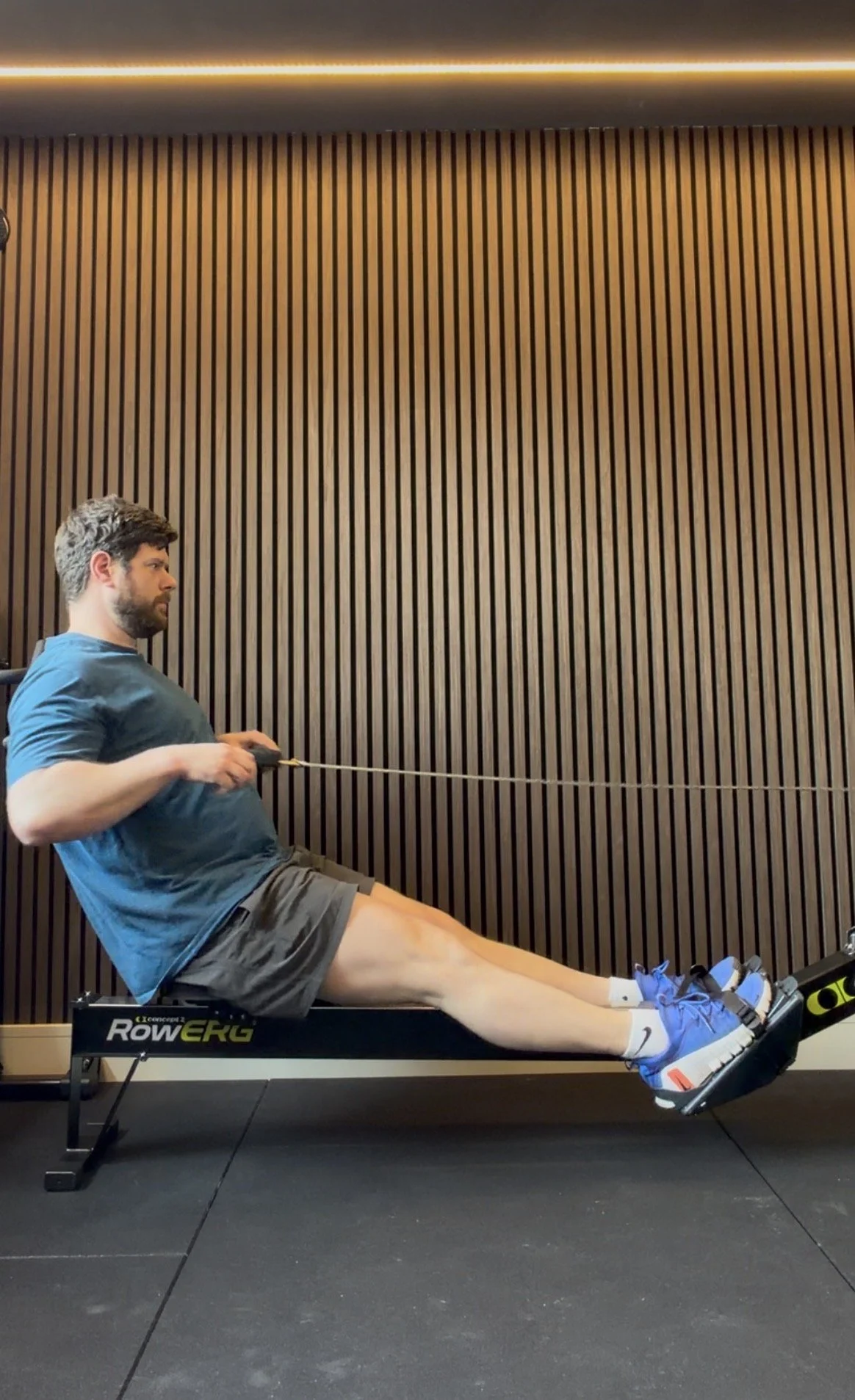 Man sitting on a workout rowing machine, pulling with a resistance band, in front of a wooden panel wall.