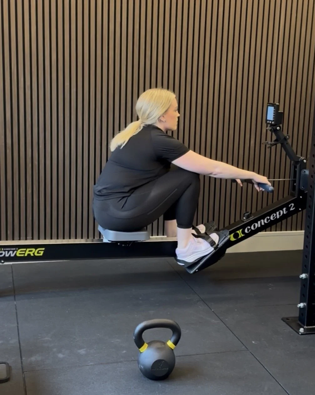 A woman with blonde hair in a ponytail working out on a rowing machine in a gym. She is sitting low, holding the handle with one arm extended, and wearing black athletic clothes and white sneakers. A kettlebell is on the floor nearby, and the background features a wooden panel wall.