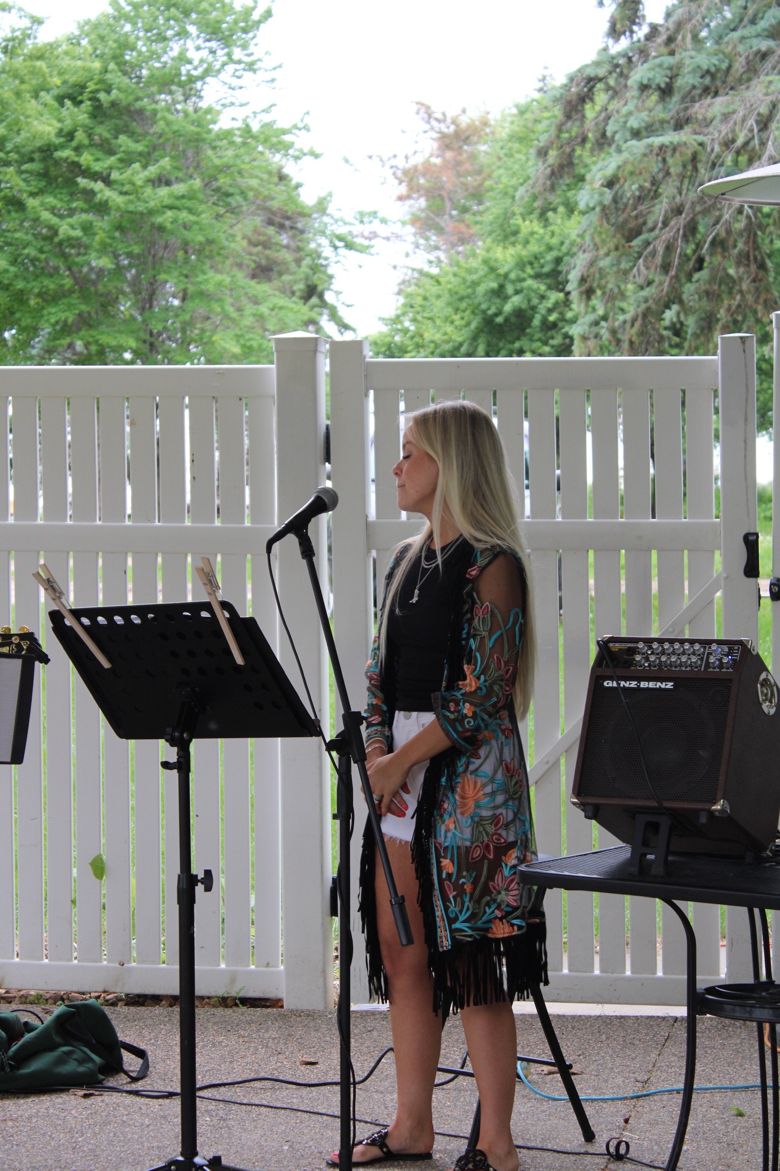 A woman with long blonde hair, wearing a black dress with colorful floral patterns and black fringe at the bottom, stands with her eyes closed in front of a microphone, possibly singing or speaking, outdoors with a white fence and green trees in the background.