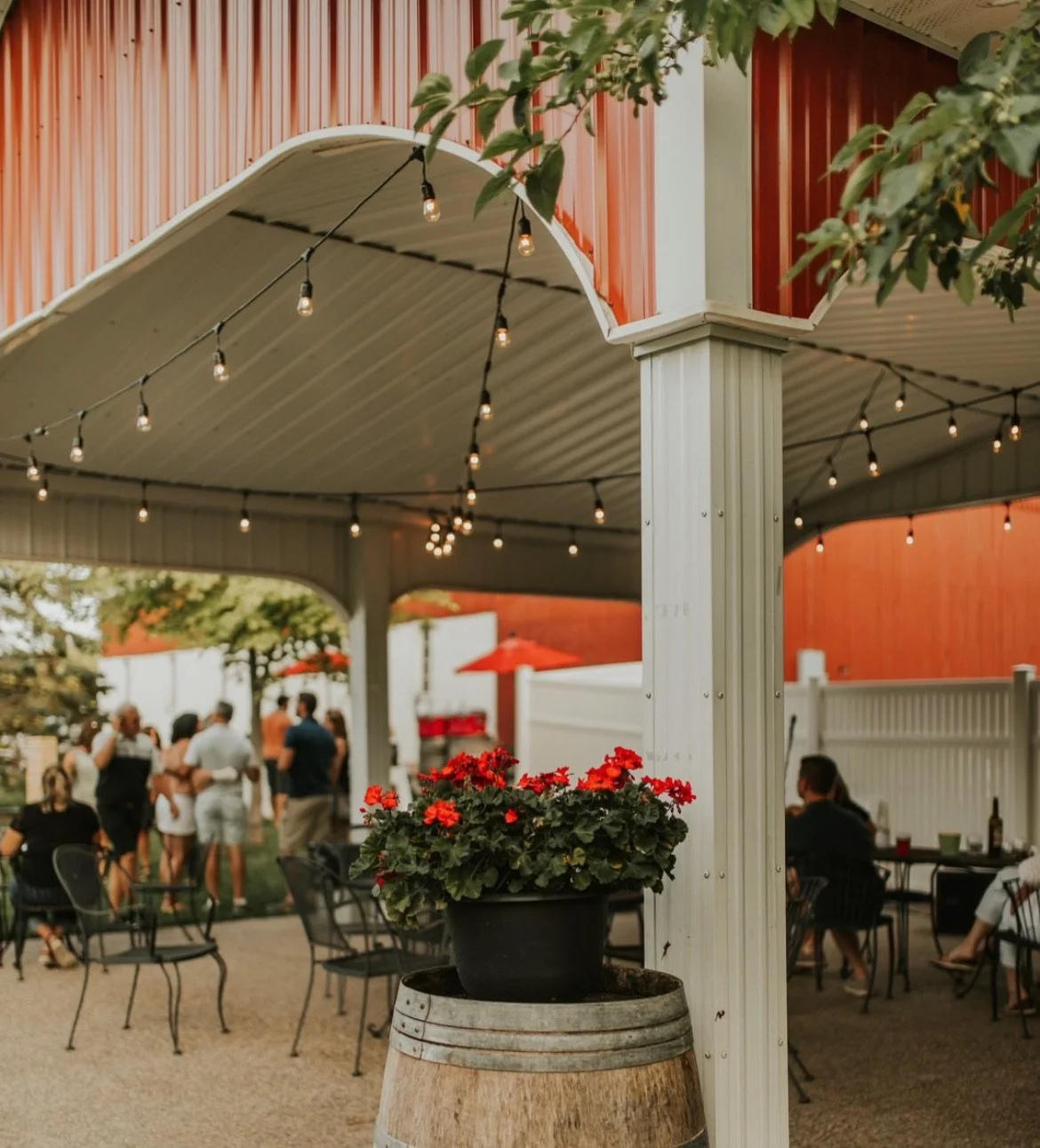 An outdoor patio area with string lights, a white fence, and a red flower planter in the foreground, with people socializing under a covered area and a building with red siding in the background.