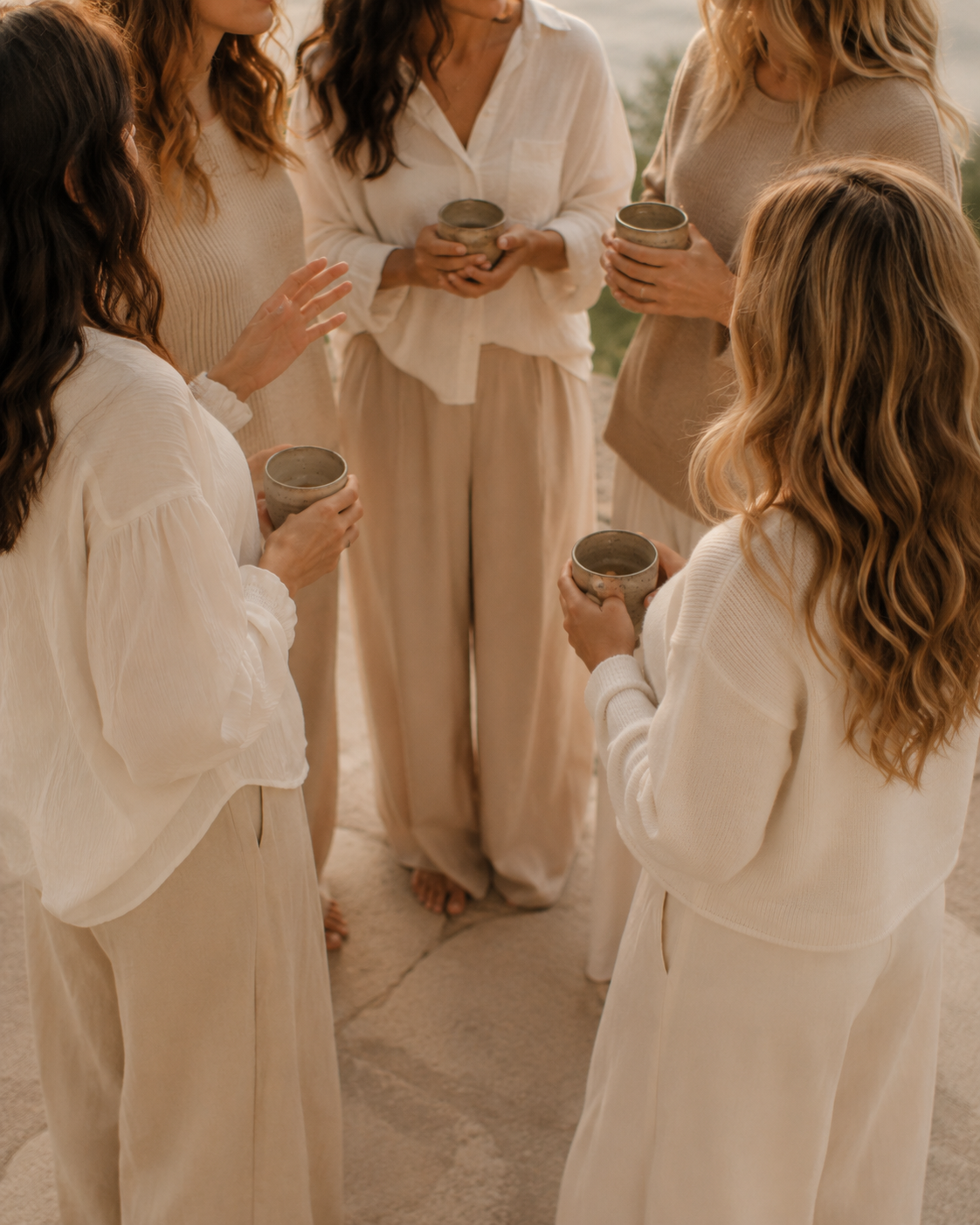 Six women standing in a circle outdoors, holding cups, engaging in conversation, dressed in neutral tones.