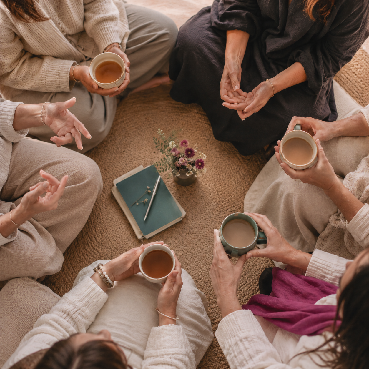 A group of women sitting in a circle on a woven rug, holding mugs of tea, with a small table with a flower vase and notebooks in the center.