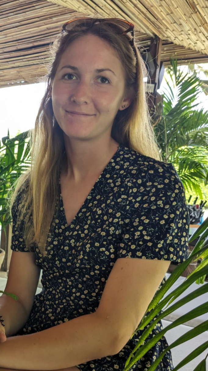 A young woman with long blonde hair, wearing sunglasses on her head, sitting outdoors at a table with greenery in the background.