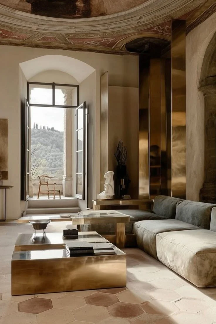Interno di un soggiorno elegante con divano beige, tavolini in specchio, una scultura bianca e un'apertura verso un balcone con vista su un paesaggio collinare. Soffitti affrescati