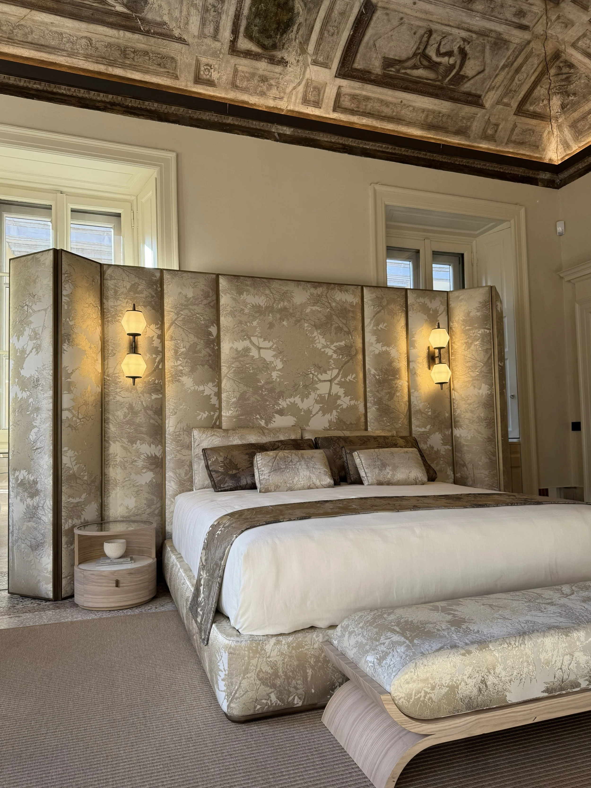 Letto matrimoniale in stile elegante con testiera, lampade a parete, divanetti coordinati, e parete divisoria decorativa in tessuto con motivo foglie.