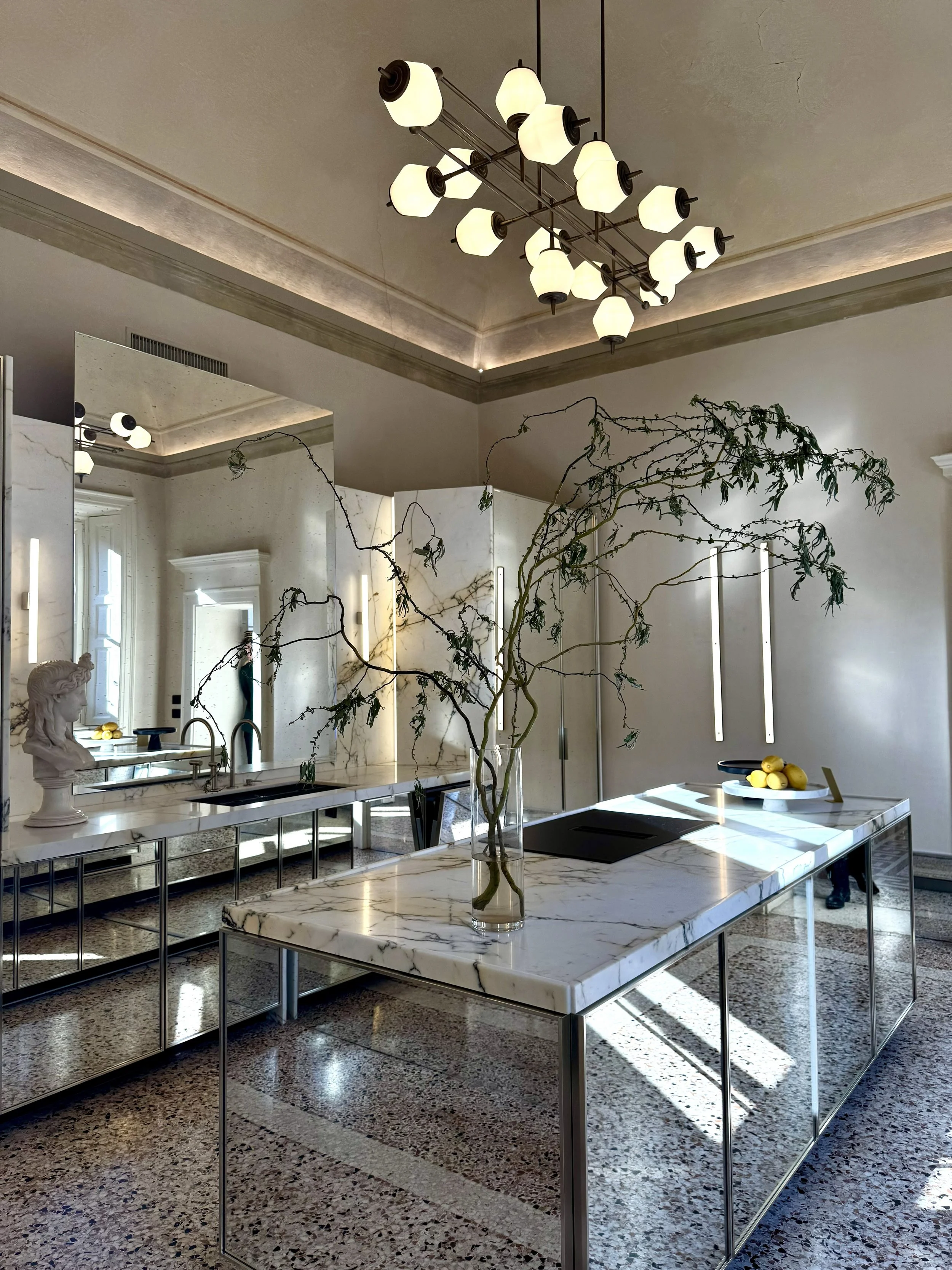 Interno di una cucina moderna con isola in specchio e piano in marmo, un lampadario presente sopra, un vaso con rami decorativi e una statua sulla pianura, pareti con specchi e luci a tiraggio verticali