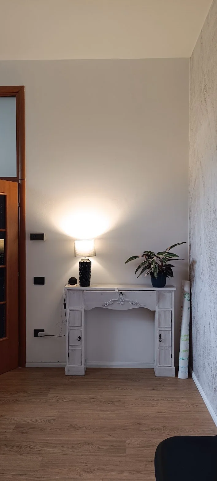 Interno di una stanza con pavimento in legno, una console bianca decorata, una lampada accesa e una pianta in vaso sul tavolo. A sinistra, una porta di legno con vetri, e a destra, un rotolo di pellicola adesiva e qualche oggetto semi nascosto.