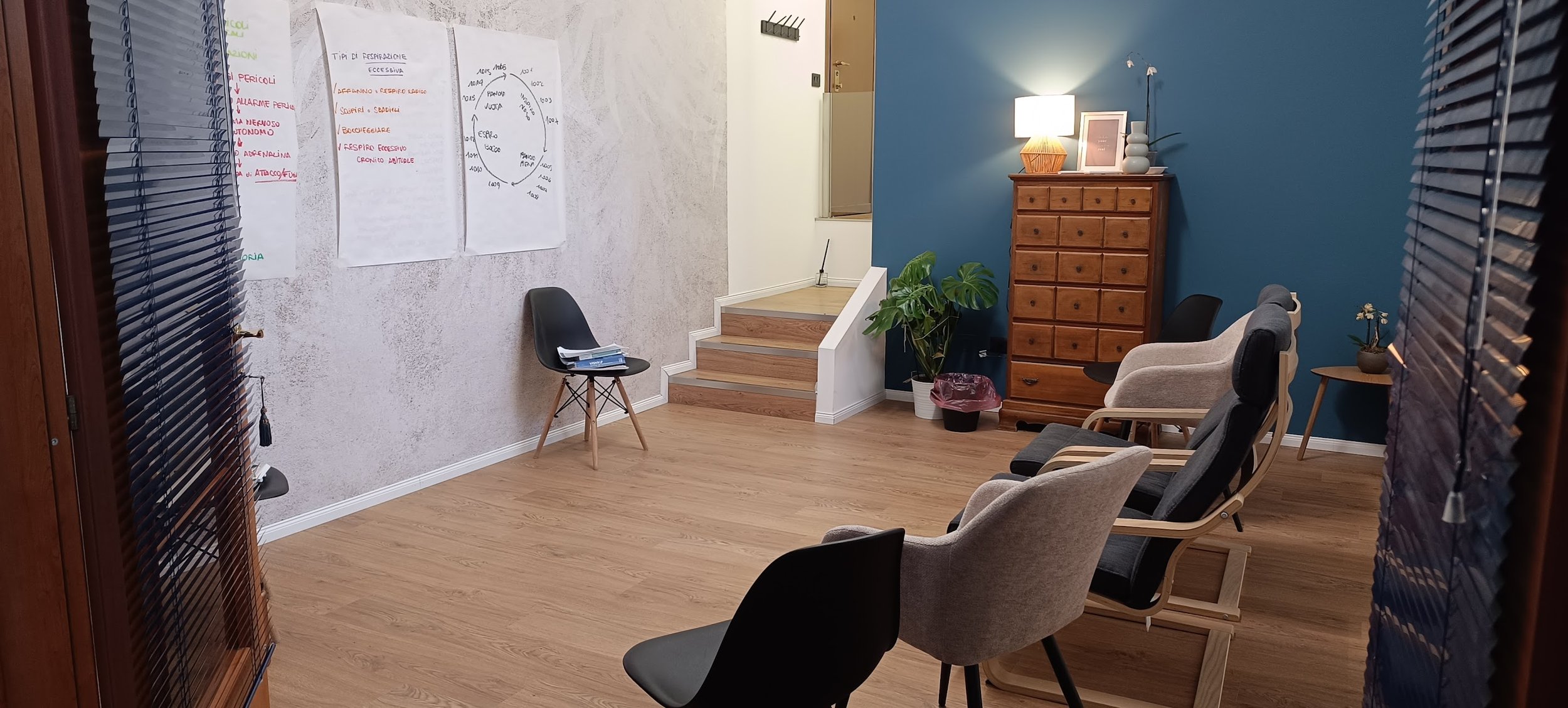 Sala riunioni con sedie di vari stili, un armadio in legno, piante, una lampada da tavolo, e tabelle appese alla parete laterale.