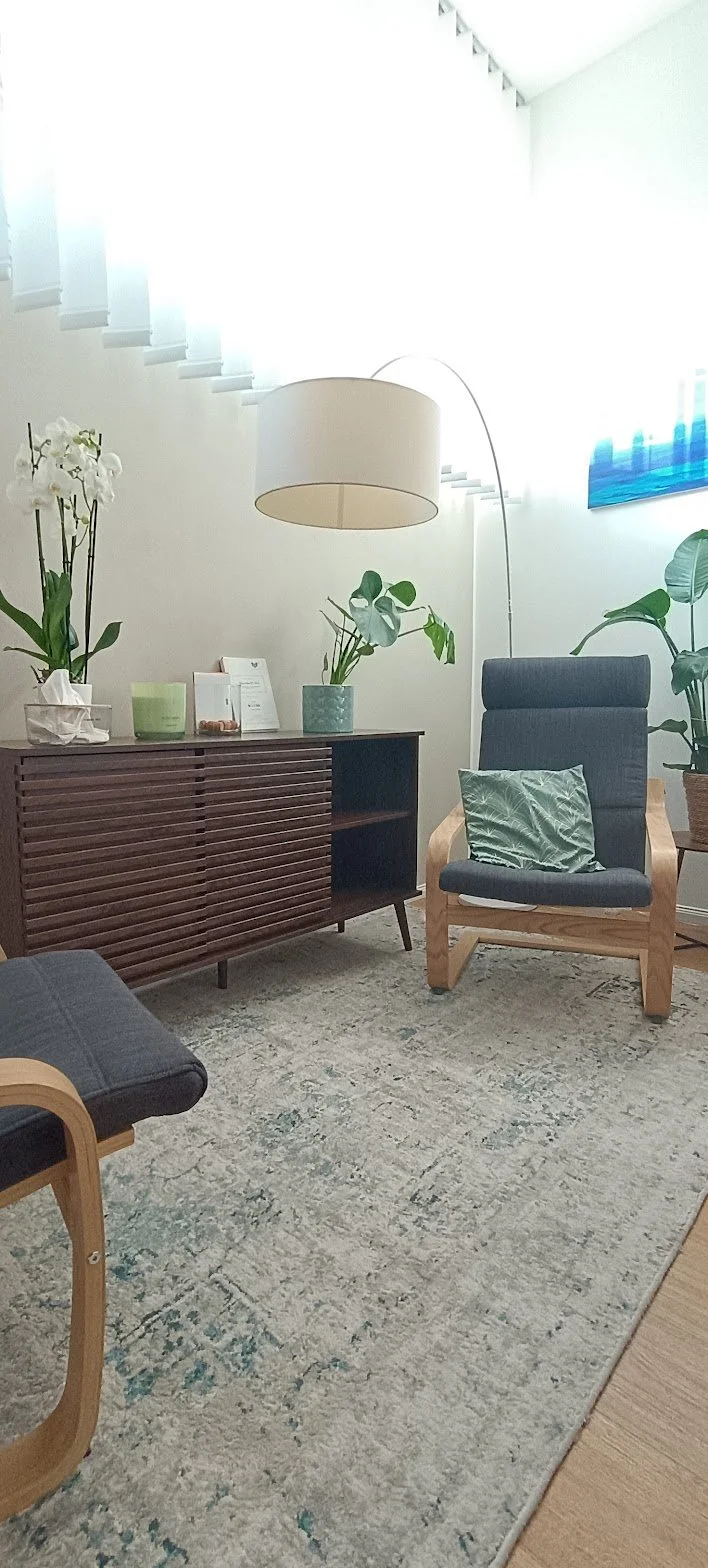 Soggiorno moderno con poltrona in legno e tessuto, bancone in legno scuro con piante verdi e oggetti decorativi, lampada a soffitto con fili sospesi, tappeto beige con dettagli verdi, pareti bianche, finestra con tende e quadro astratto blu.