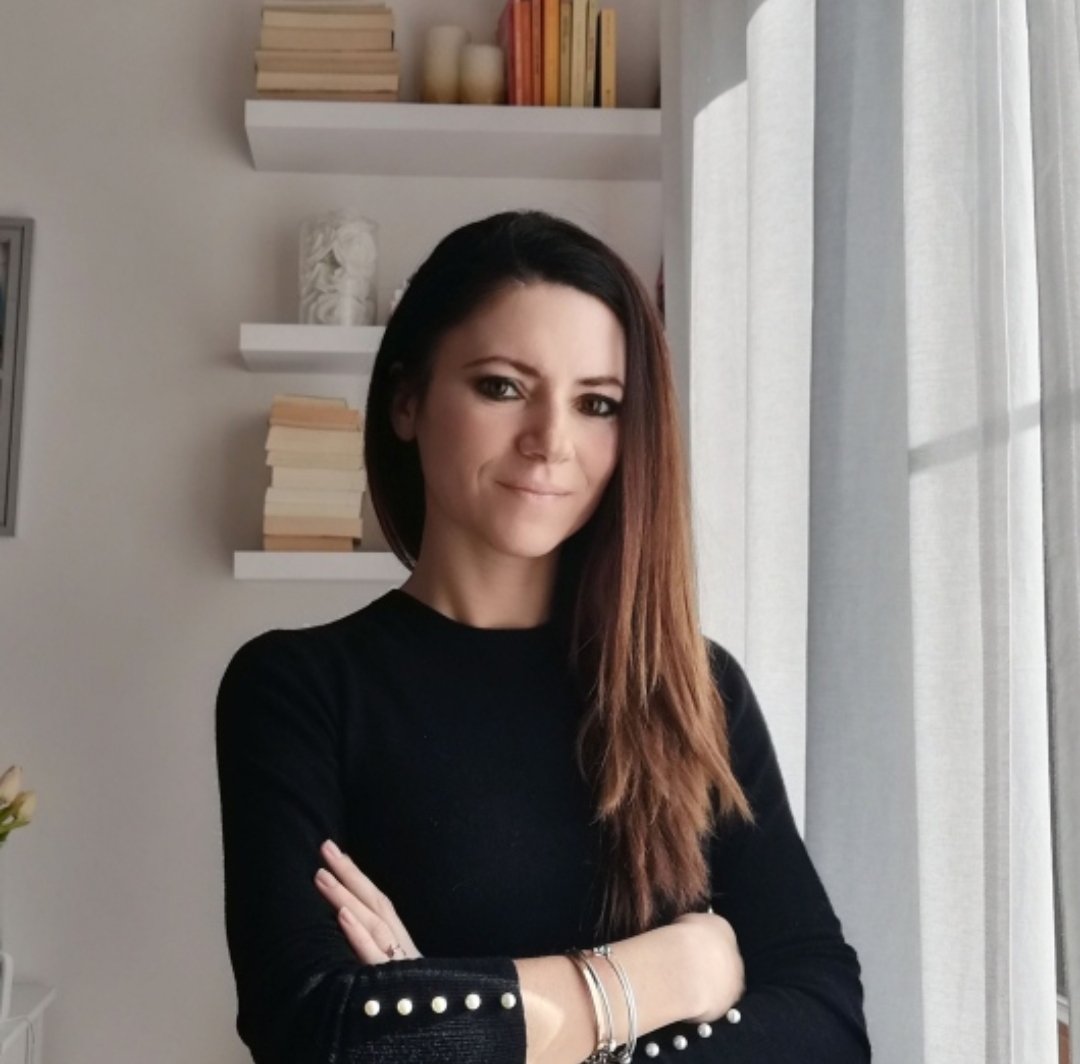 Una donna con capelli castani e vestita con una maglietta nera si trova in una stanza vicino a una finestra con tende chiare, sfondo con scaffali bianchi pieni di libri e decorazioni.