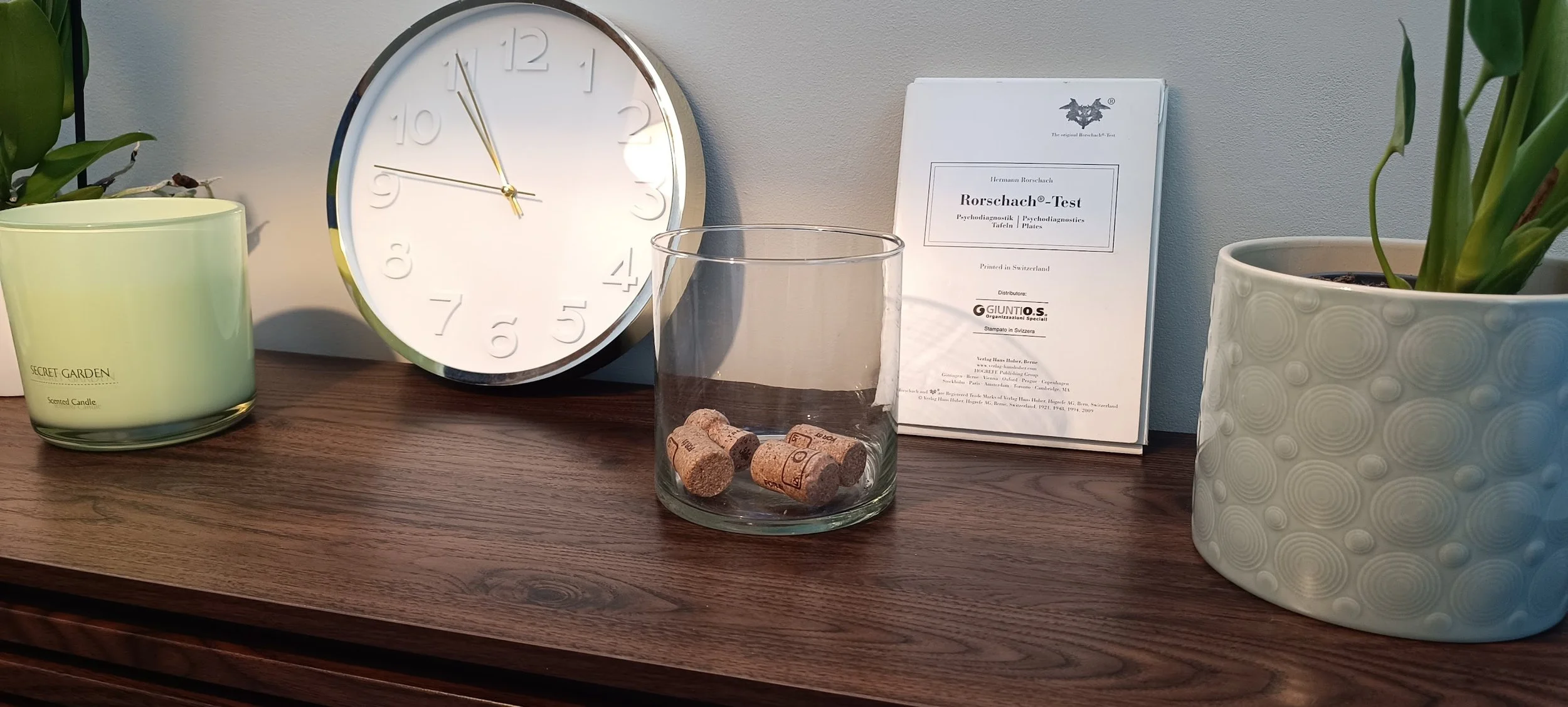 Oggetti sulla scrivania: orologio da parete, due piante in vaso, candela profumata, bicchiere di vetro con tappi di sughero e libreria con libro sulla psicodiagnostica Rorschach.