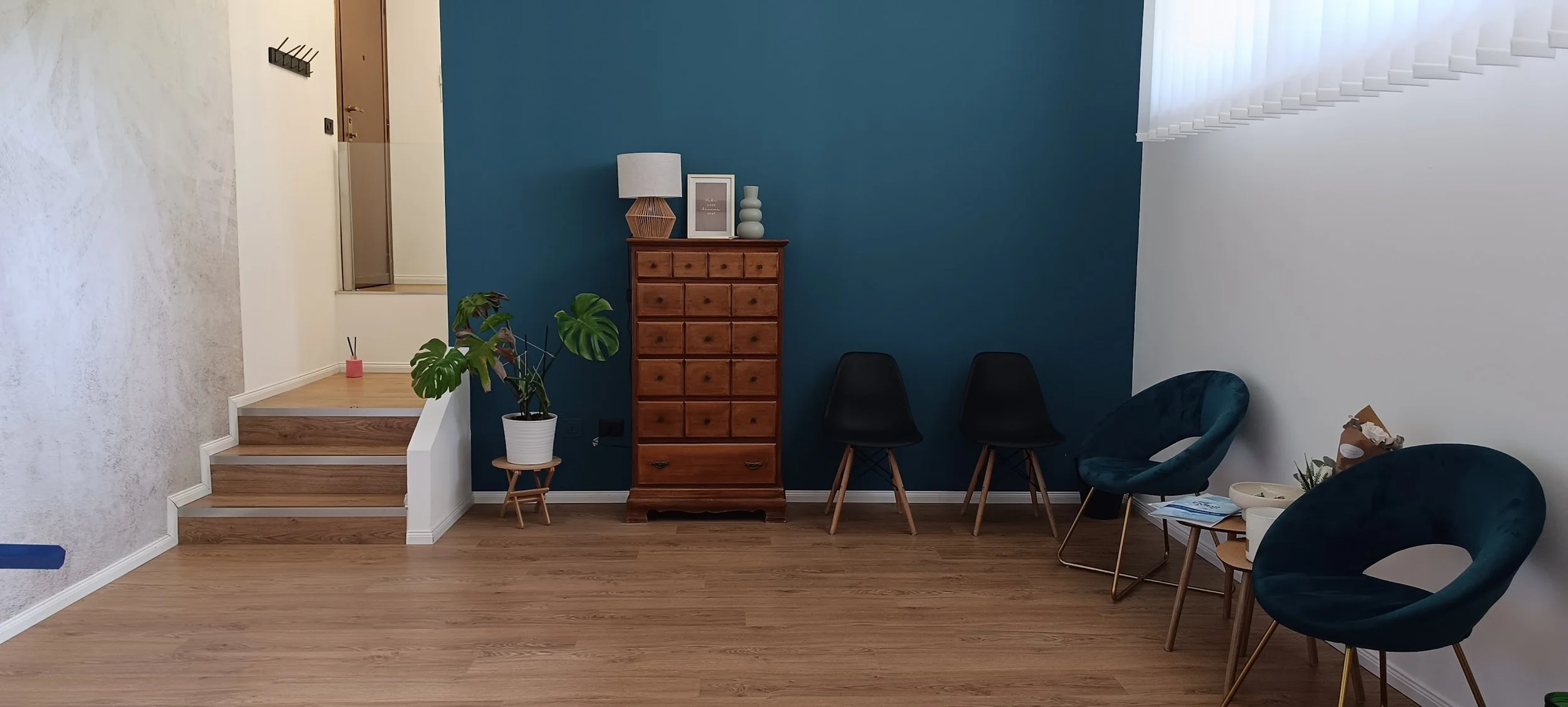 Interno di un soggiorno con parete blu, panca di legno, piante in vaso, sedie nere e rivestimenti in legno, illuminato dalla luce naturale.