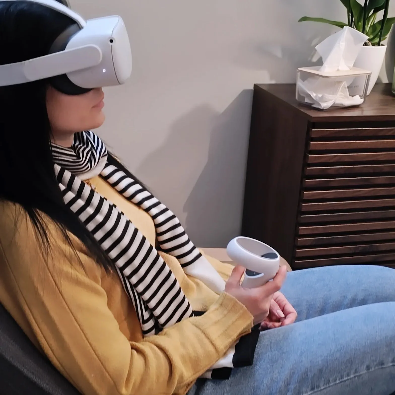 Donna con maglietta a righe nere e bianche e giacca beige, seduta su una poltrona, indossa un visore di realtà virtuale e tiene in mano un controller.