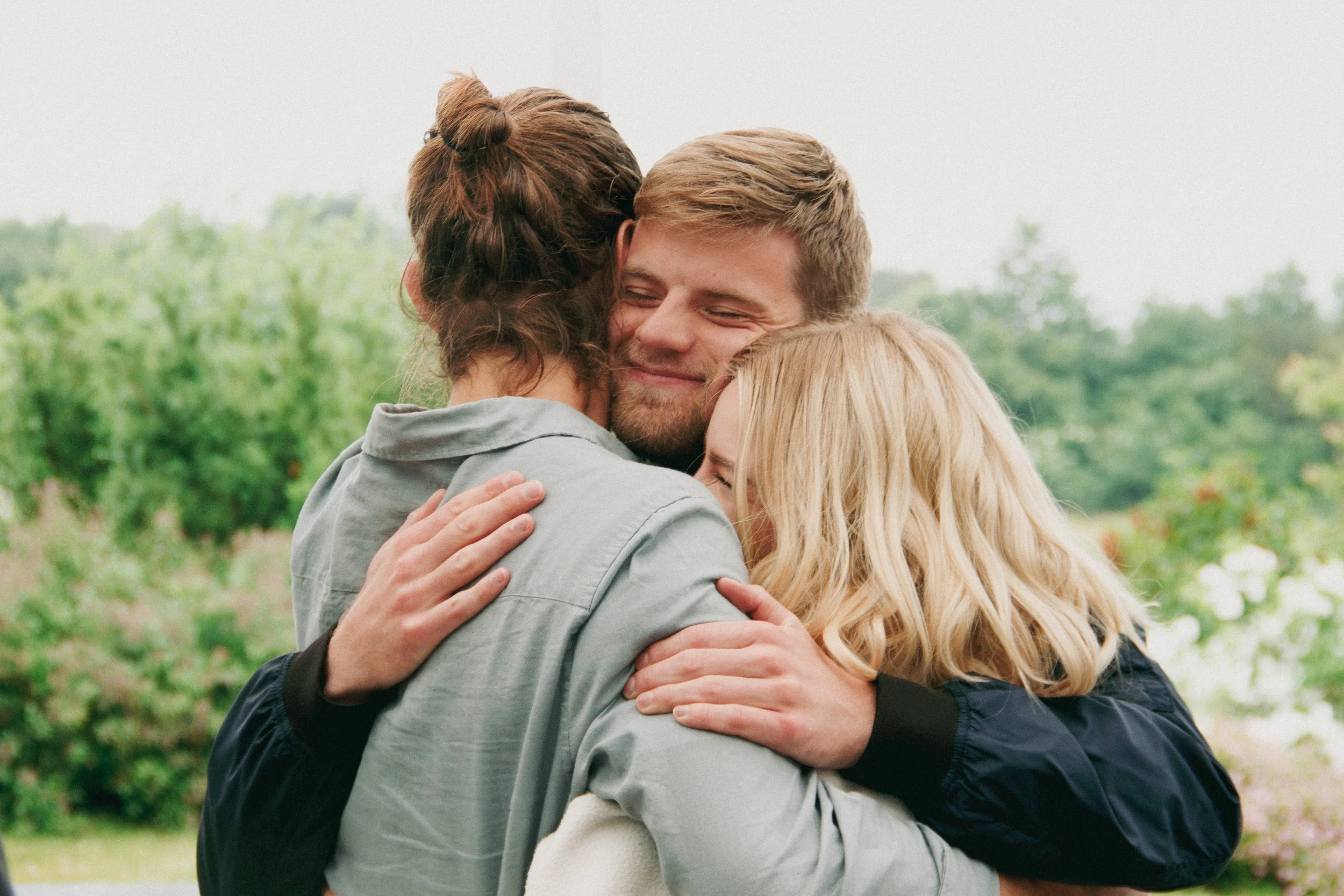 Tre personer, to kvinder og en mand, omfavner hinanden og smiler i et udendørs område med grønne træer i baggrunden.