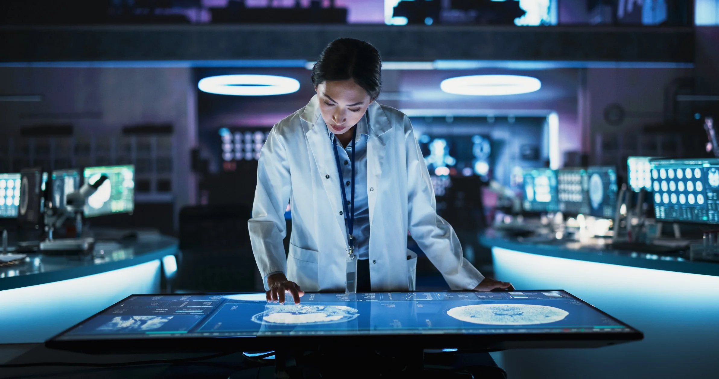 A researcher in a white lab coat studying data on a large illuminated touchscreen table in a dark control room.