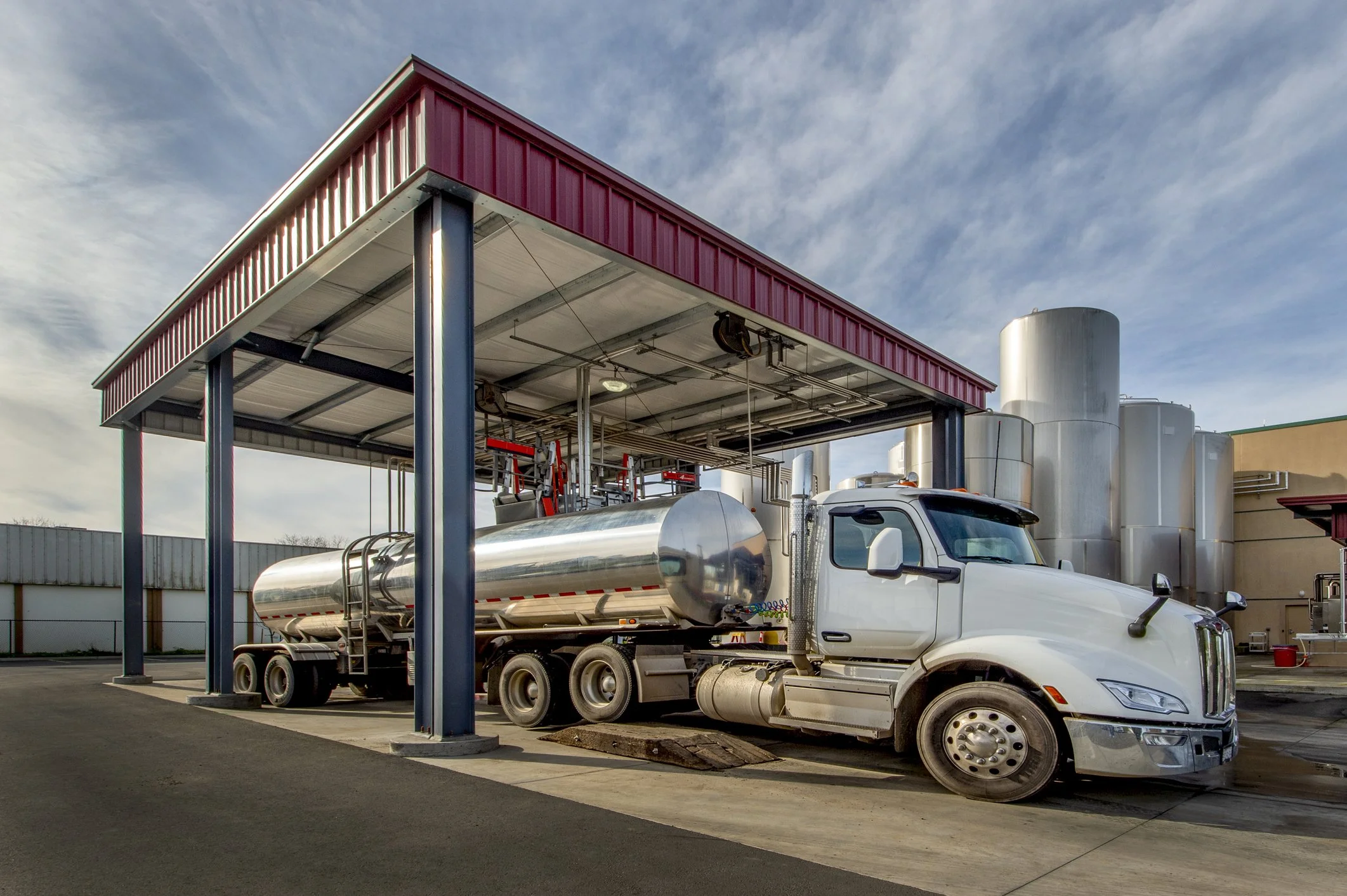 A tanker truck parked under a loading canopy at a fuel and lubricants terminal, storage tanks behind.