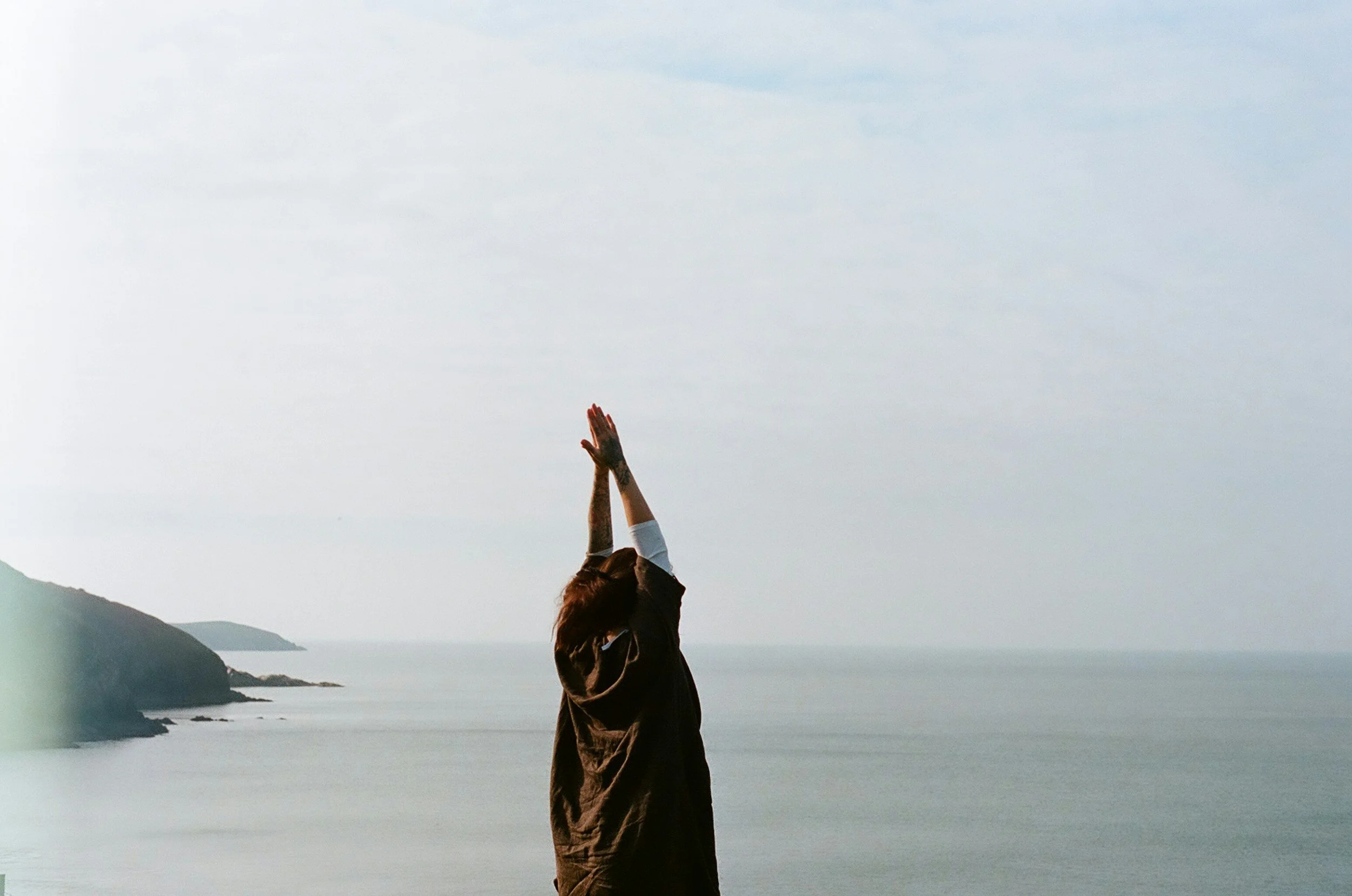 A person with long hair wearing a dark jacket standing outdoors near water, reaching up with both hands; the background features mountains and a cloudy sky.