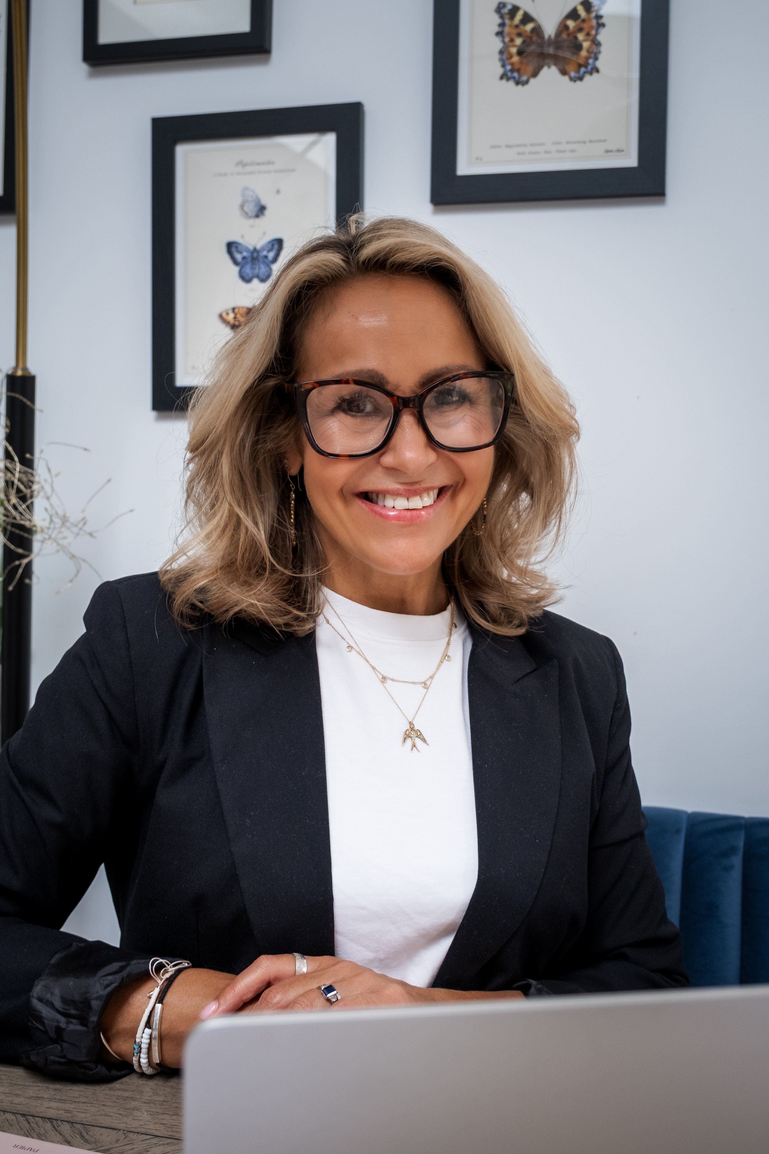 A woman with blonde wavy hair and glasses smiling at the camera, sitting at a desk with her hands clasped in front of her. She is wearing a black blazer and a white top, accessorized with layered necklaces and earrings. Behind her, there are framed butterfly posters on the wall and a white wall in an office or conference room setting.