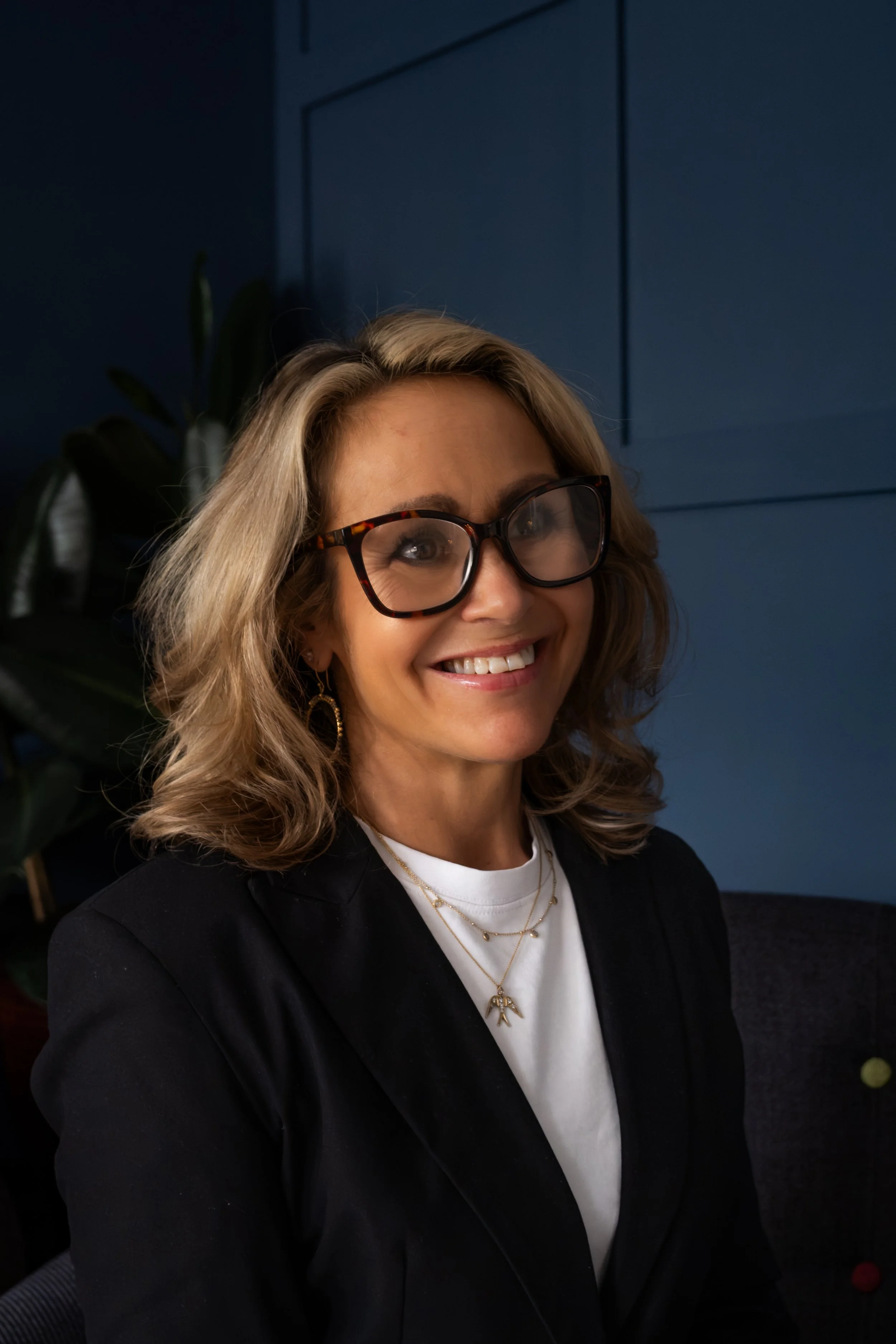 A woman with blonde, wavy hair wearing black glasses, gold earrings, a layered gold necklace, a white shirt, and a black blazer, smiling indoors with a dark blue wall and a potted plant in the background.