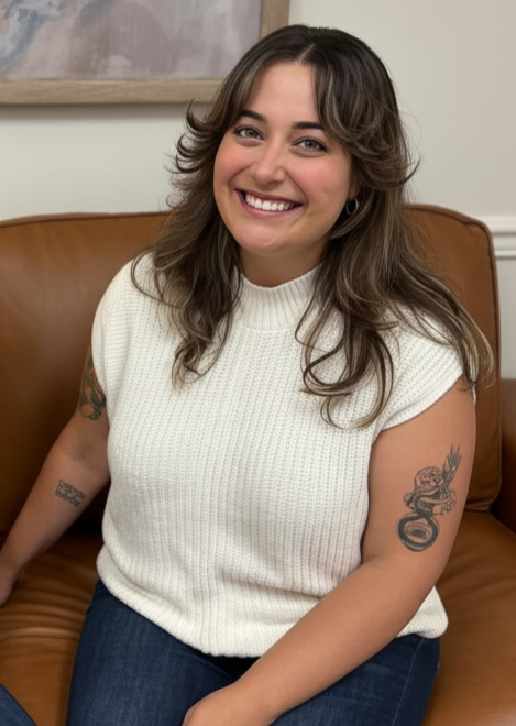 A smiling woman with long wavy hair, tattoos on both arms, wearing a cream knitted sleeveless top, sitting on a brown leather couch indoors.
