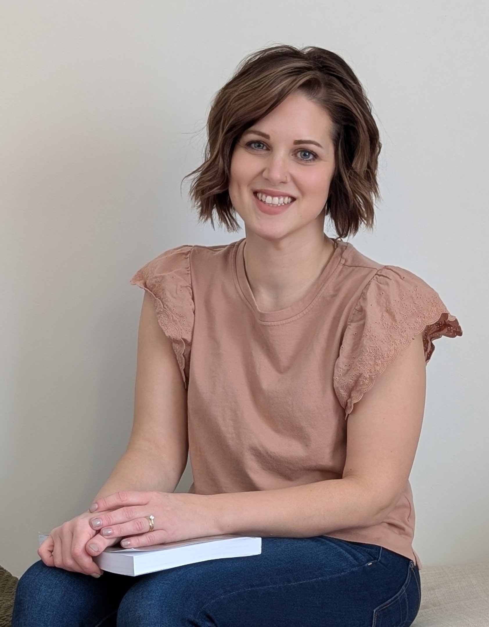 A woman with short, wavy brown hair and blue eyes smiling while sitting cross-legged with a book on her lap, wearing a light pink blouse with lace details on the sleeves, against a plain light-colored wall background.