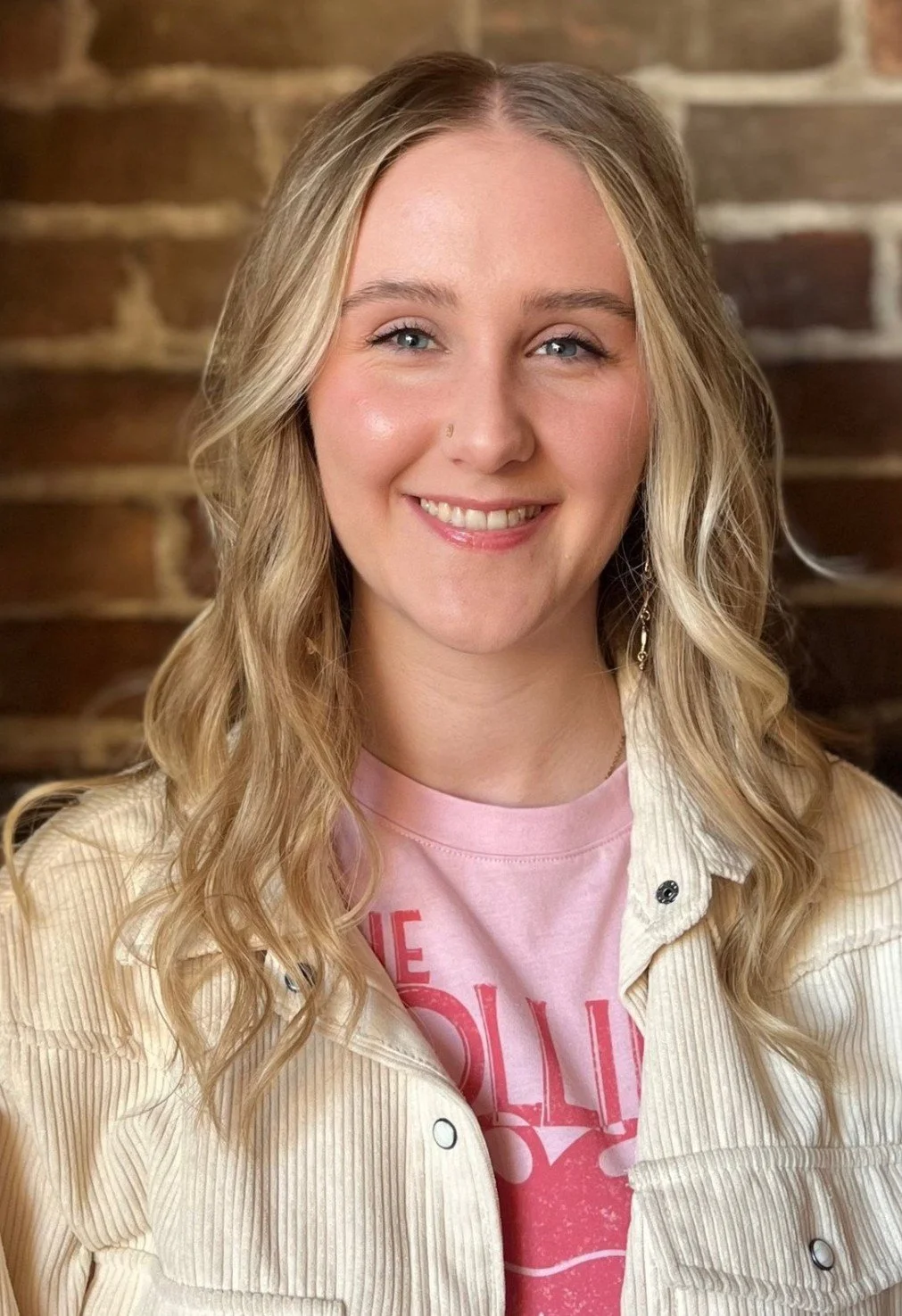 A young woman with long curly blonde hair, smiling at the camera, wearing a pink graphic t-shirt and a cream-colored corduroy jacket, standing in front of a brick wall.